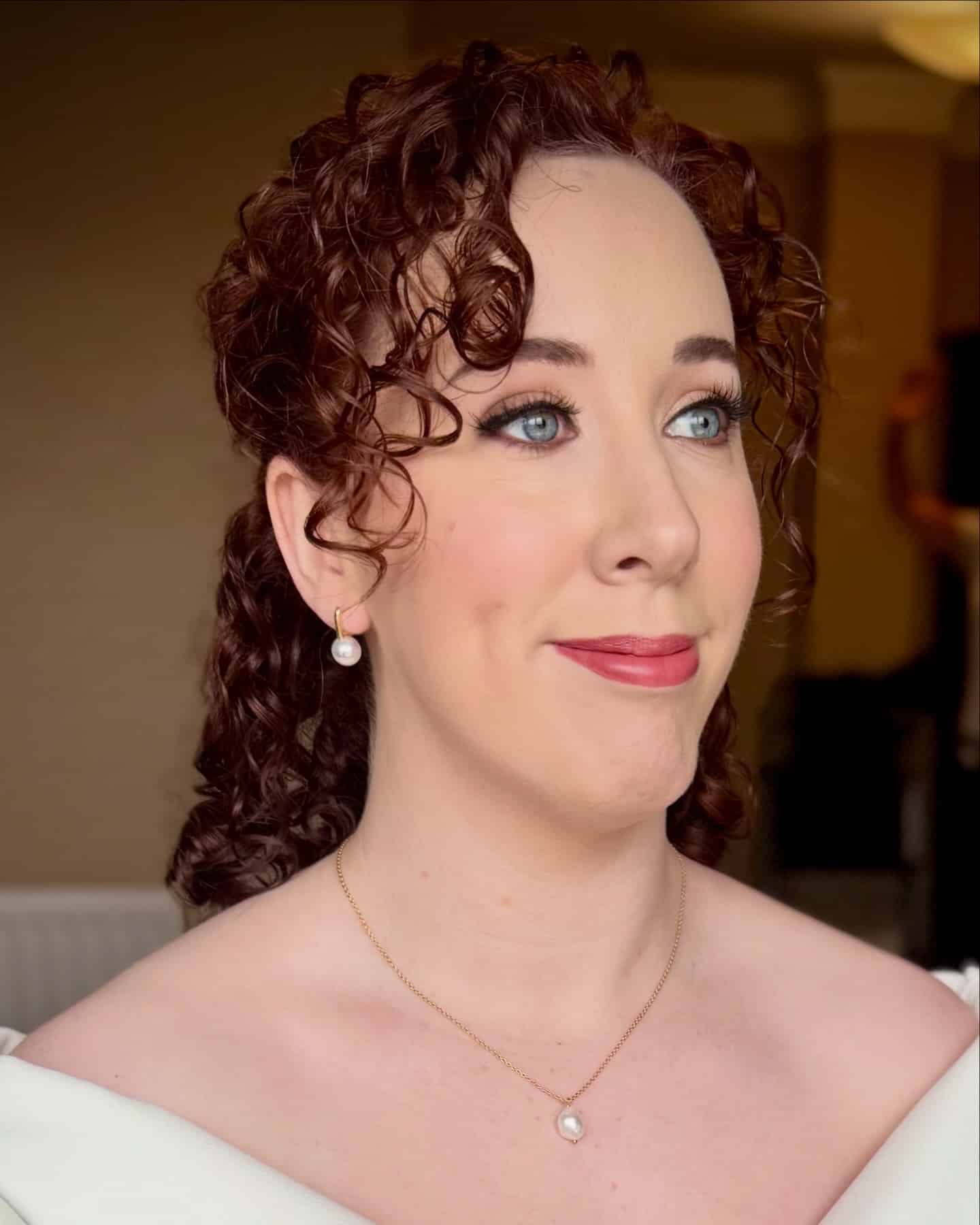 Woman with curly hair wearing pearl jewelry and a white top, smiling softly indoors.