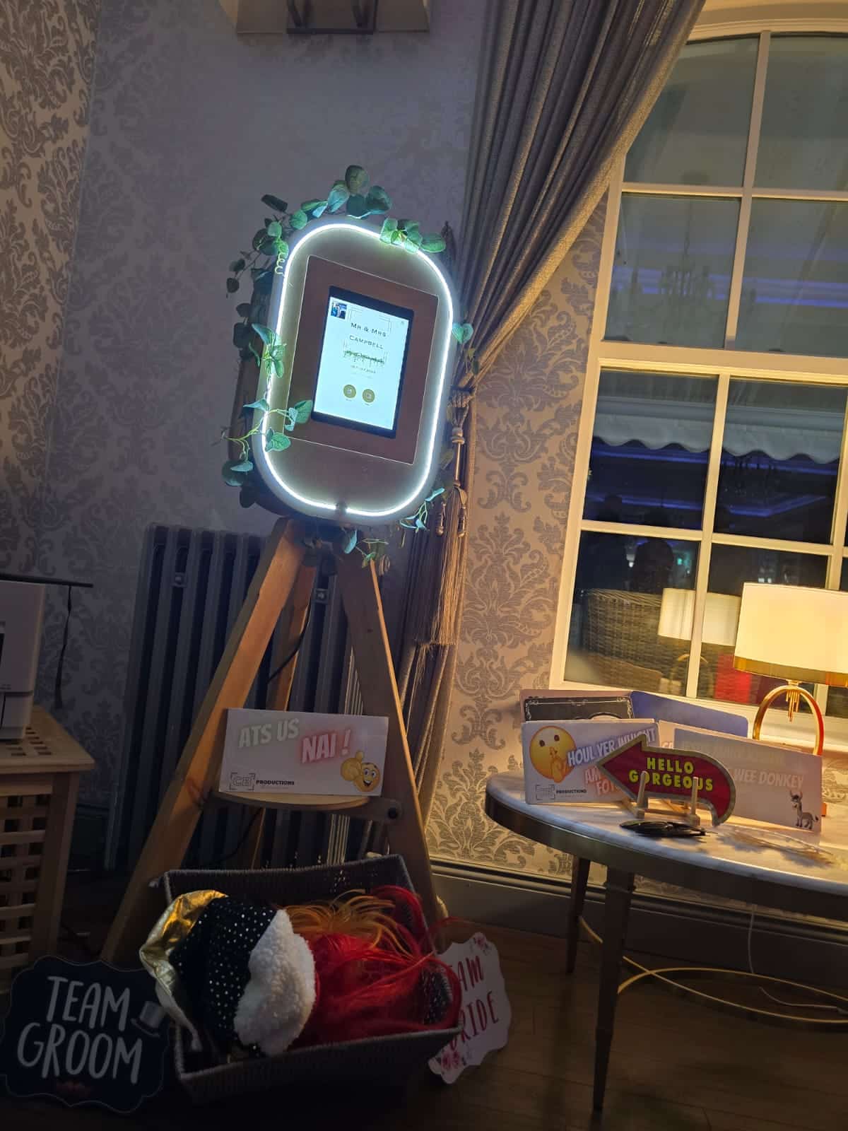 Photo booth setup with illuminated frame and props, including "Team Groom" and "Team Bride" signs, in a decorated room.