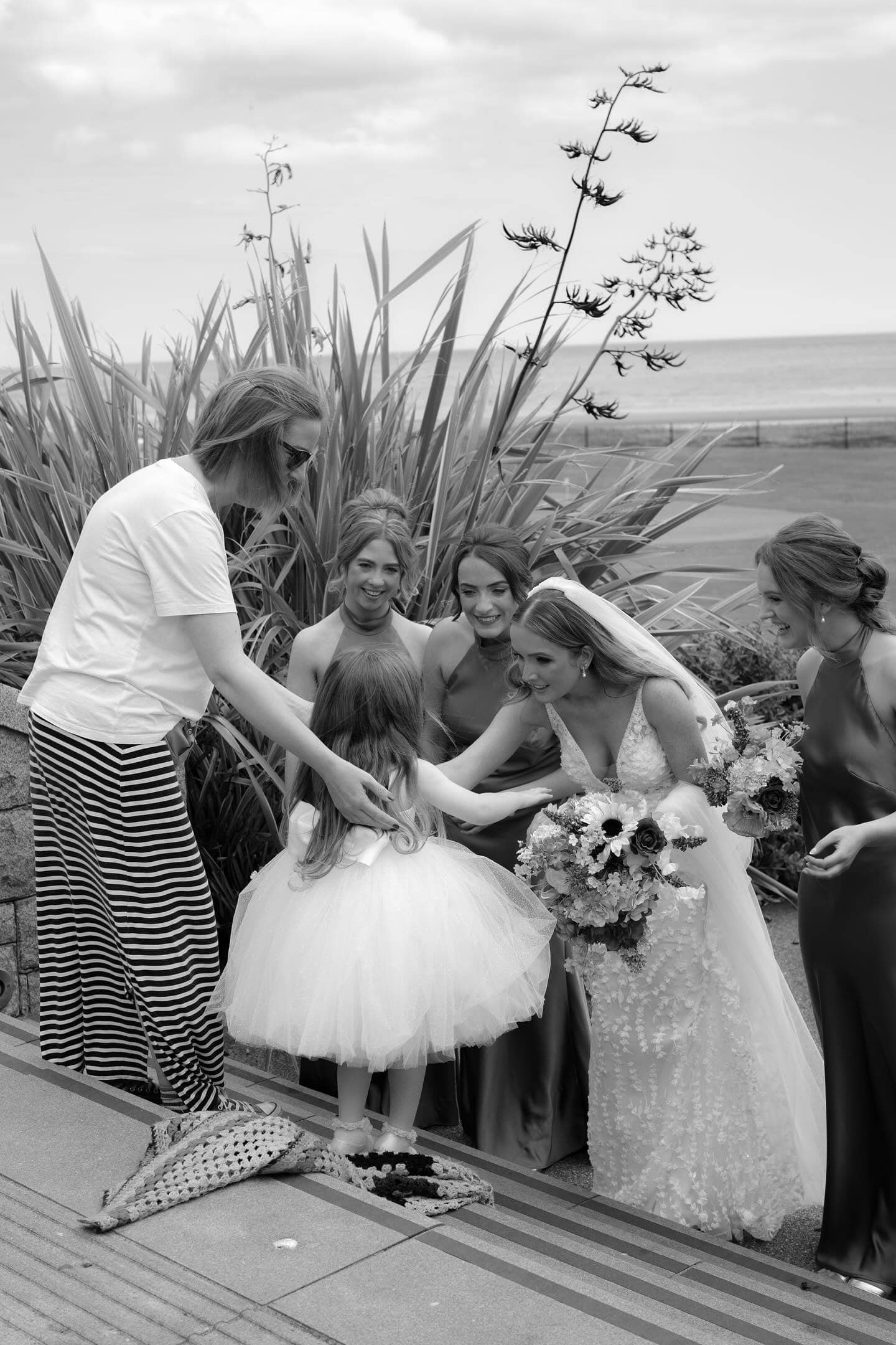 Bride and bridesmaids with a flower girl in a white dress, outdoors by the sea.
