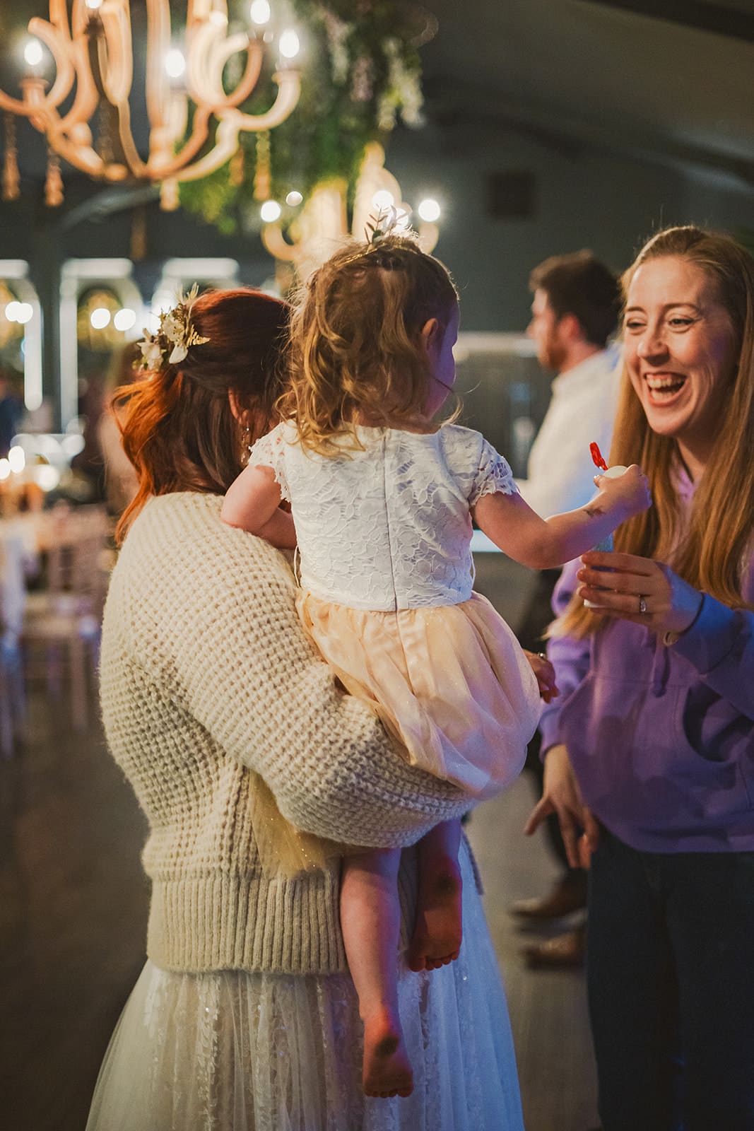 Woman holding a child in a dress at an indoor event, with another woman smiling nearby.