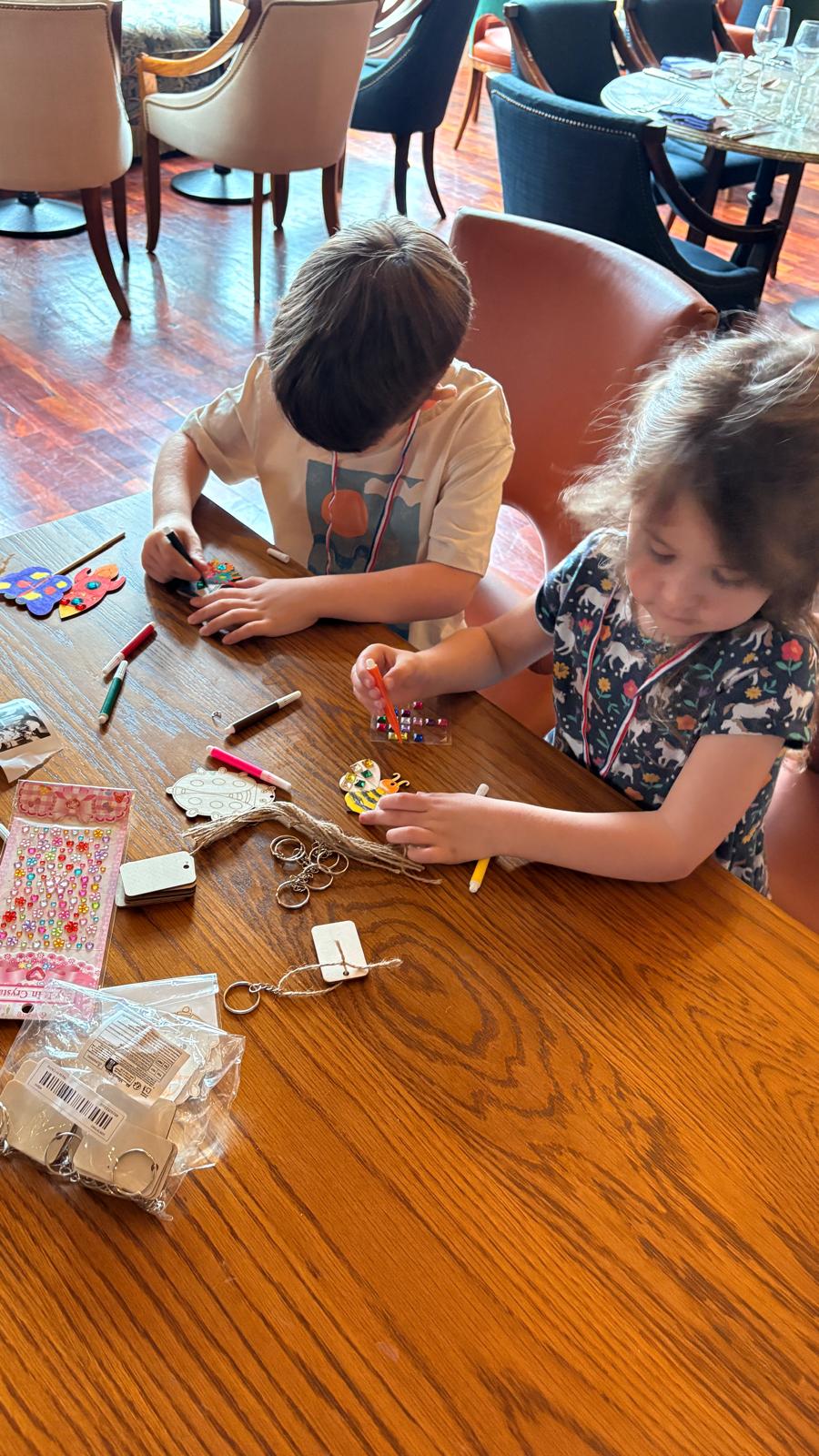 Children crafting with markers and stickers at a wooden table, surrounded by art supplies and keychain materials.