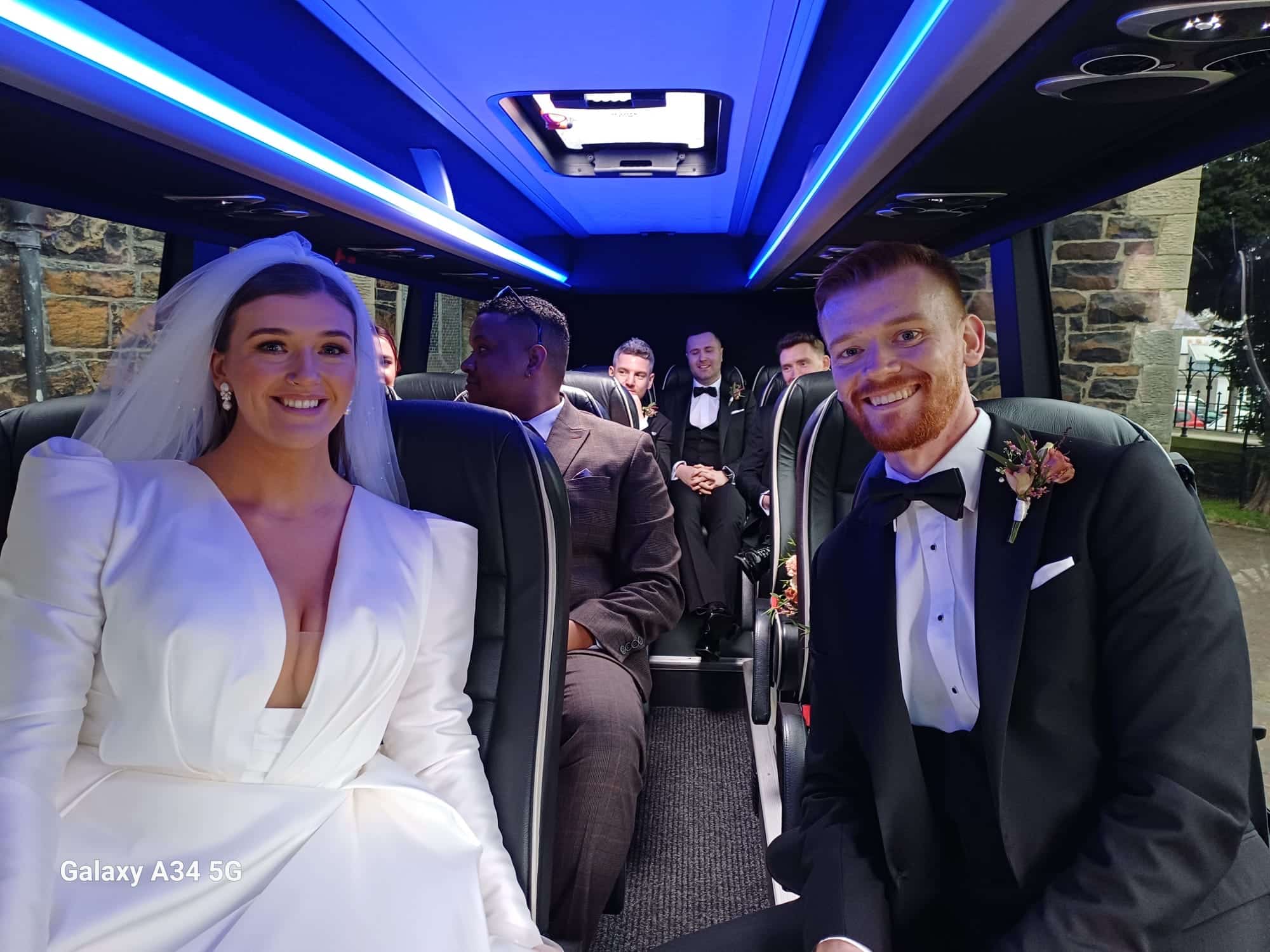 Bride and groom smiling in a limousine with friends in the background.
