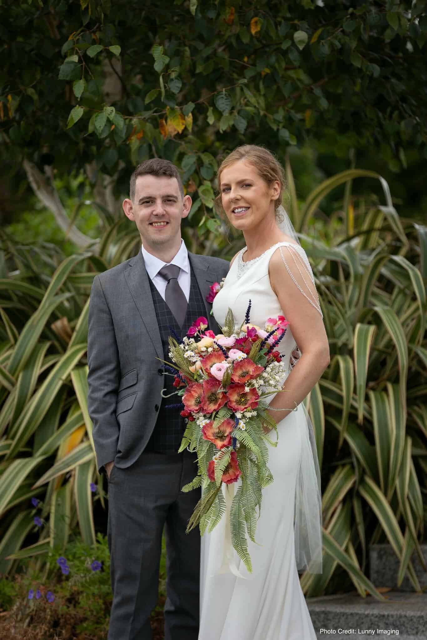 Bride holding vibrant bouquet stands beside groom in grey suit outdoors.