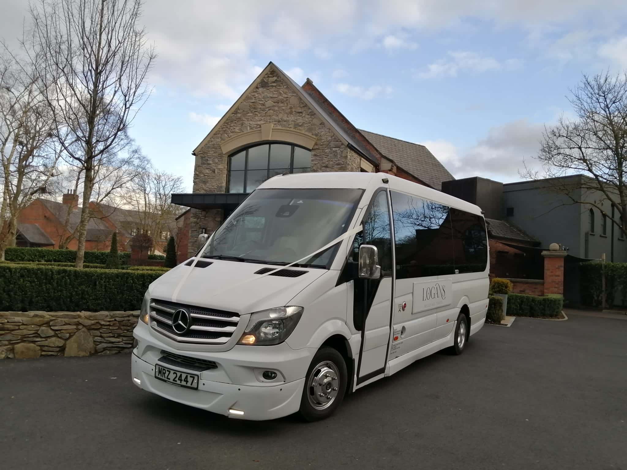 White Mercedes-Benz minibus parked in front of a stone building on a cloudy day.