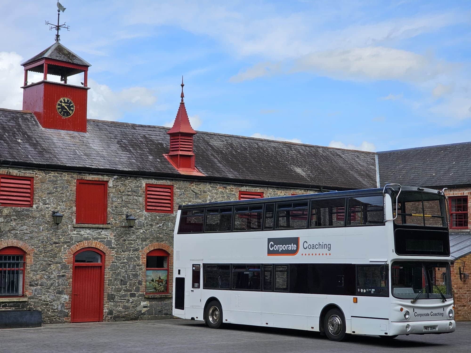 White double-decker bus labeled "Corporate Coaching" parked by a rustic stone building with red accents.