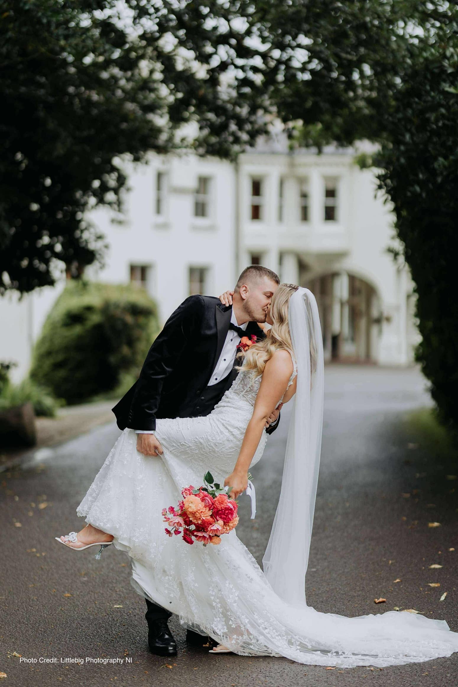 Bride and groom kissing, groom lifting bride in wedding attire, holding bouquet, on a tree-lined path.