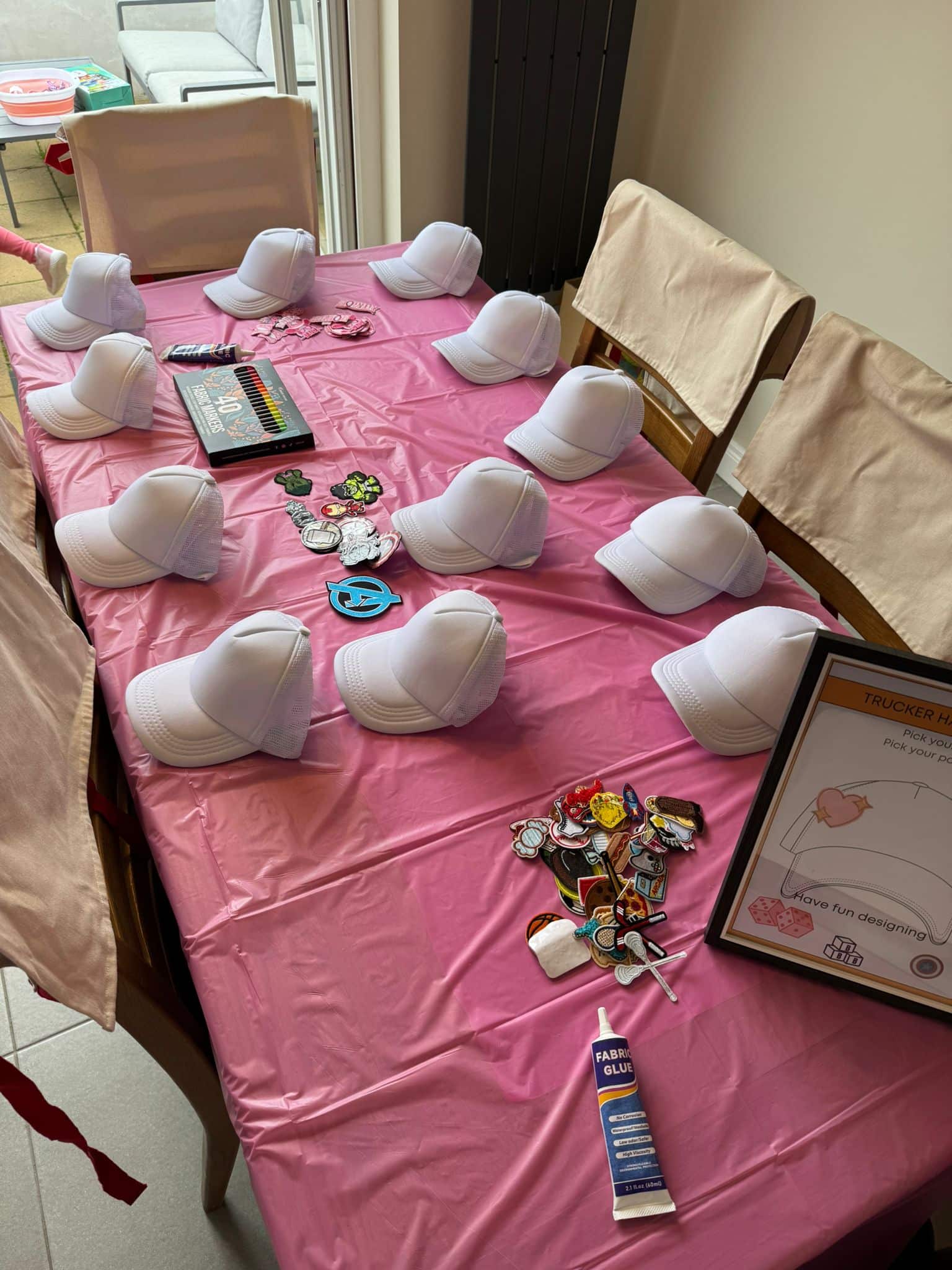 White trucker hats on a pink table with patches, markers, and fabric glue for a DIY decoration activity.