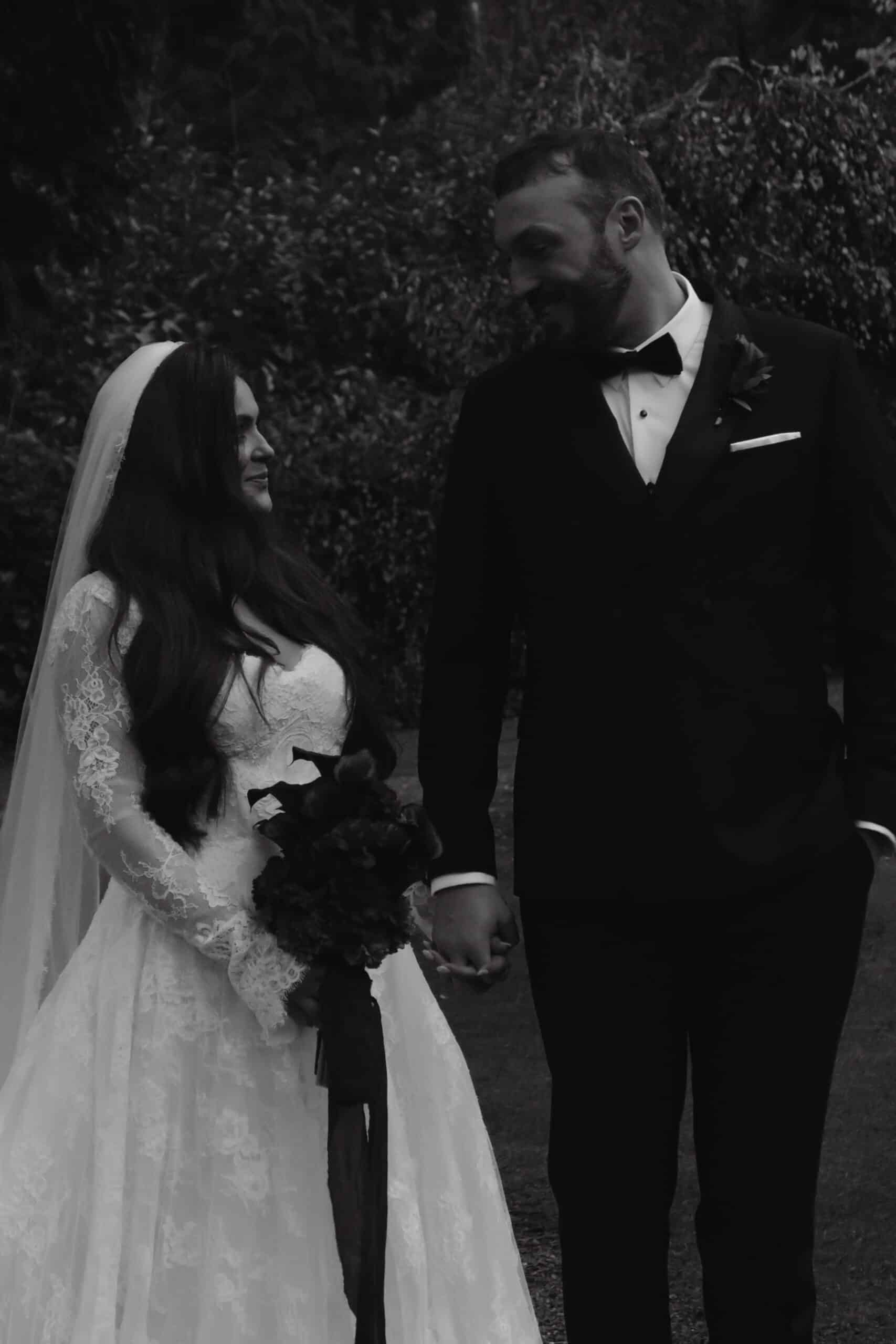 Bride and groom holding hands, both wearing formal wedding attire, standing outdoors in a wooded area.