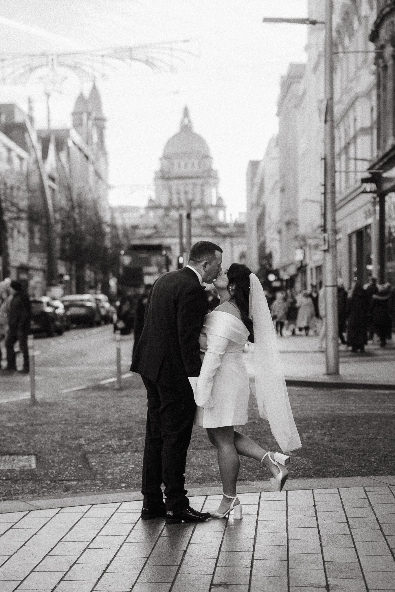 A couple kissing on a city street in black and white, with a historic building in the background.