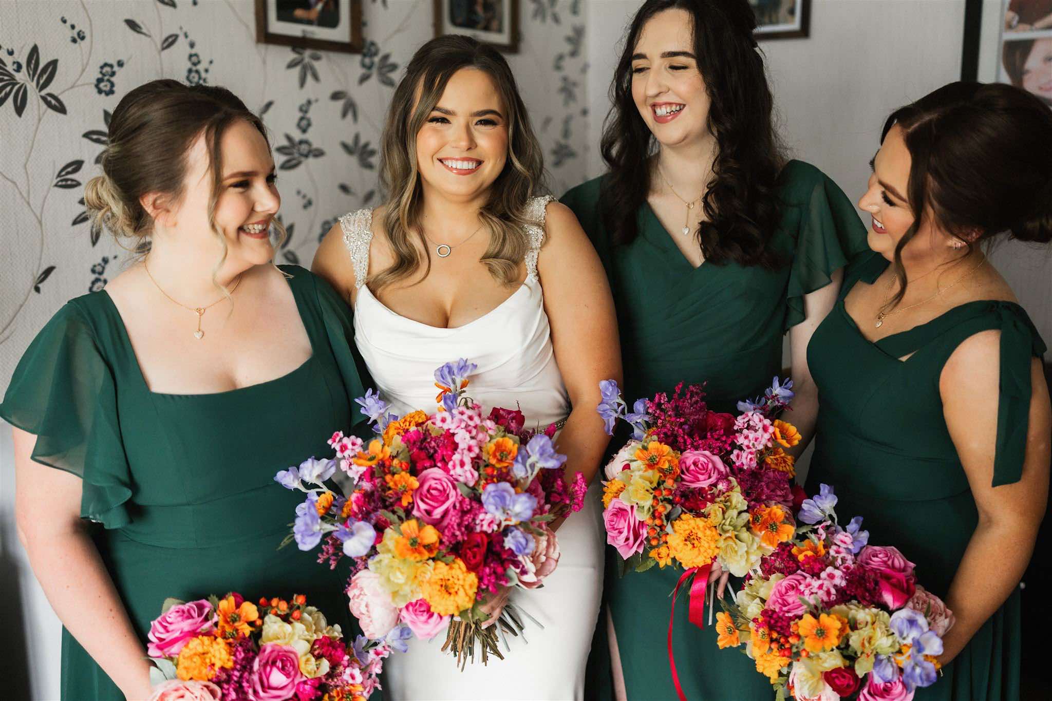 Bride and bridesmaids in green dresses holding colorful bouquets, smiling indoors.