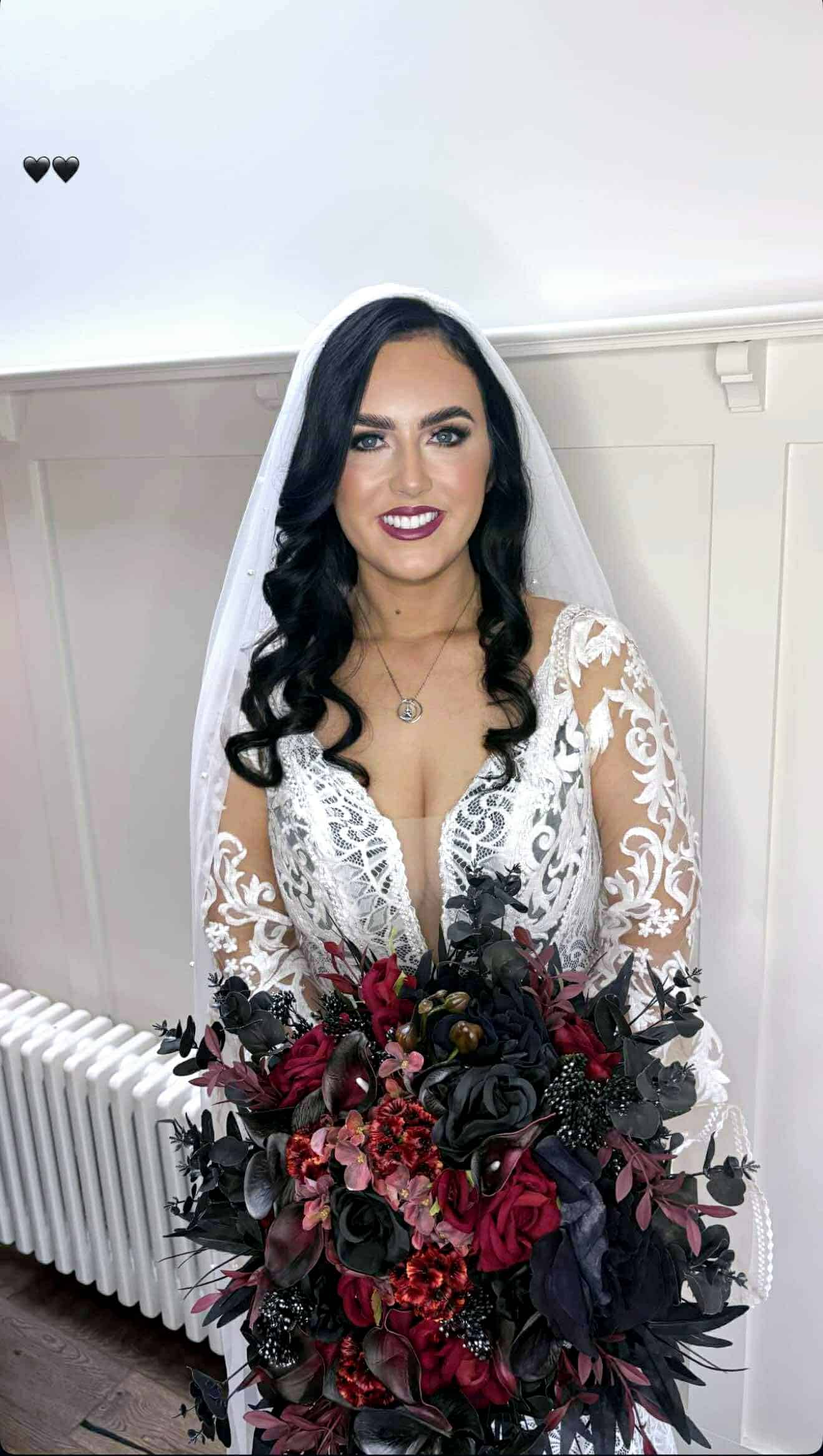Bride in lacy white dress holding a bouquet of dark and red flowers, smiling indoors.