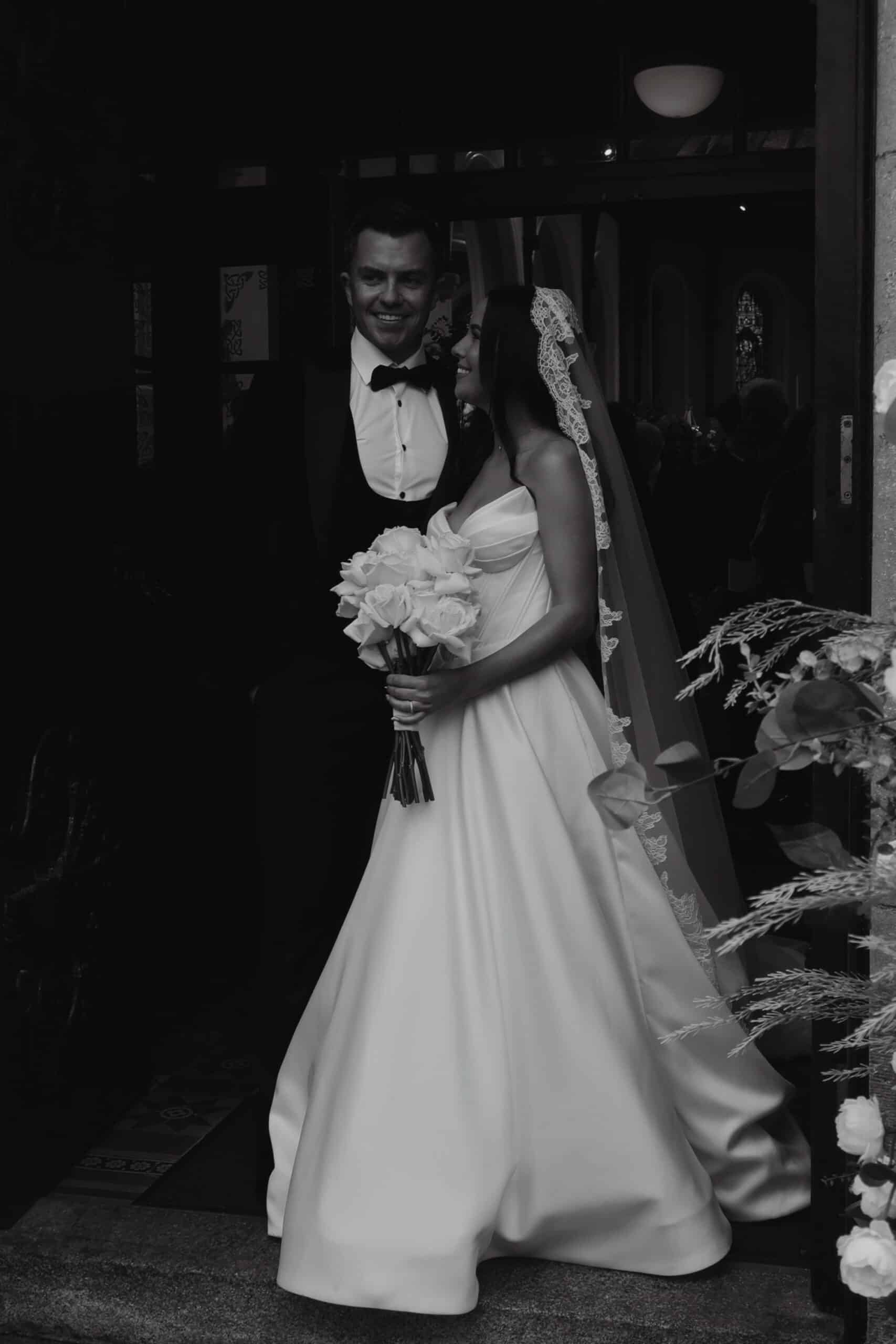Bride and groom smiling at each other, standing in a doorway, black and white photo.