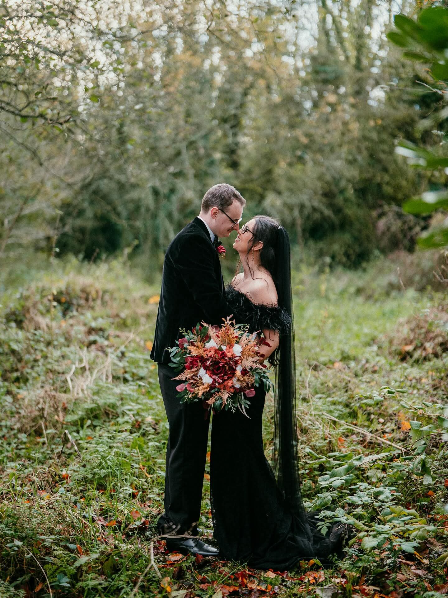 A couple in formal attire embraces in a forest, with the woman holding a bouquet of autumn flowers.