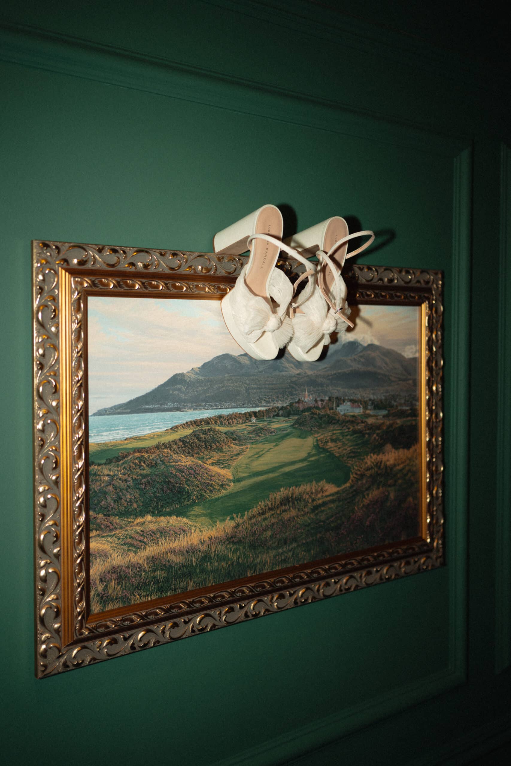 White shoes hanging on a framed landscape painting with a green wall background.
