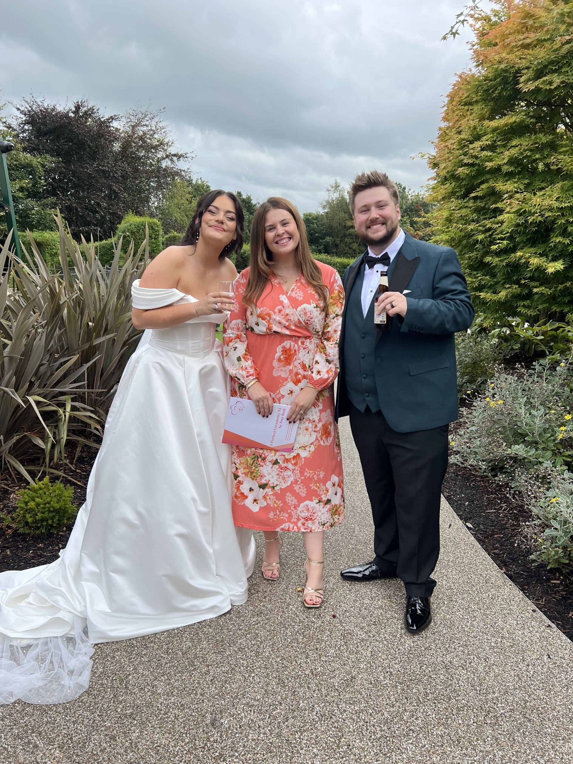Three people posing together outdoors; a bride, a woman in a floral dress, and a man in a dark suit with a green jacket.