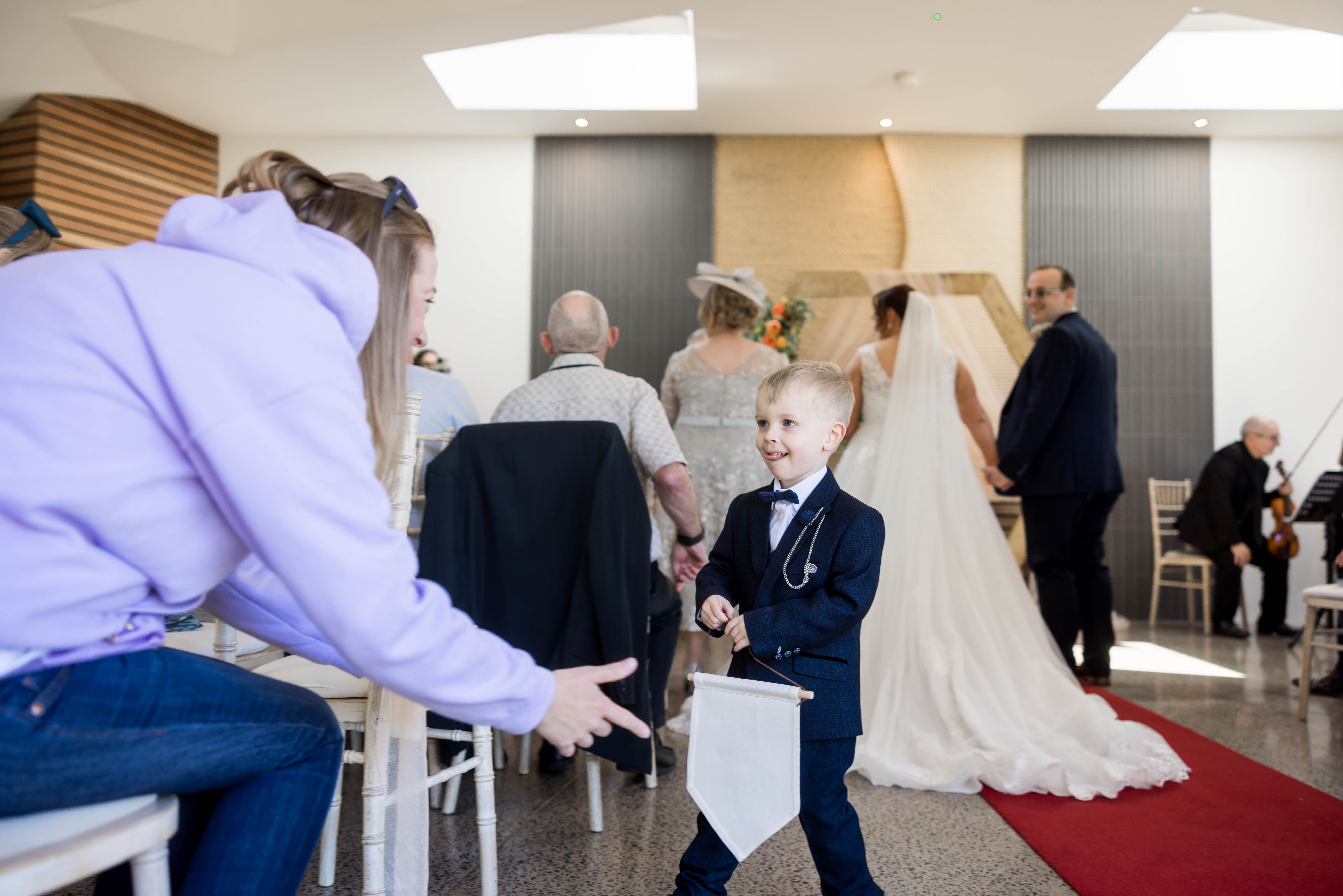 Young boy in a suit holds a banner at a wedding ceremony, with guests seated and a couple at the altar in the background.