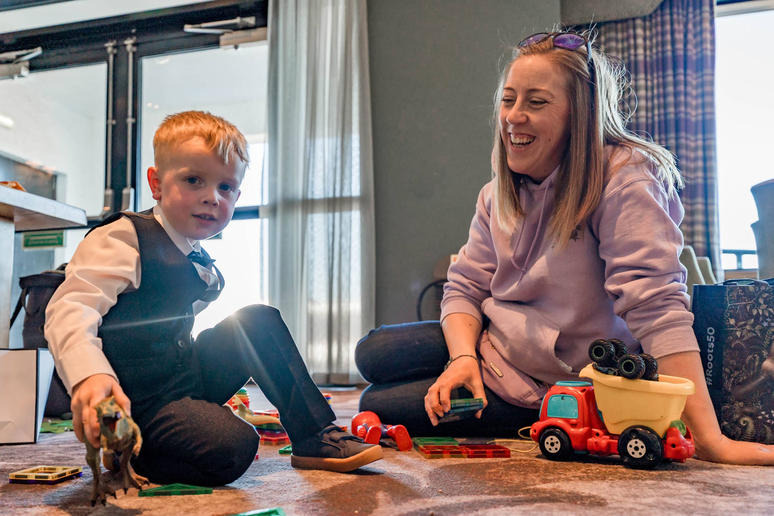 Child in formal attire playing on the floor with toys, while an adult in a hoodie smiles beside them.