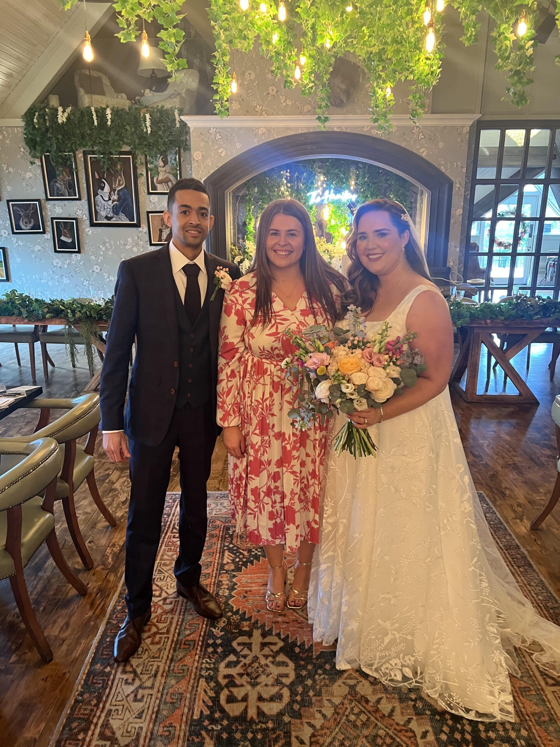 A bride in a white dress holding a bouquet poses with two guests in a decorated indoor venue with string lights and greenery.