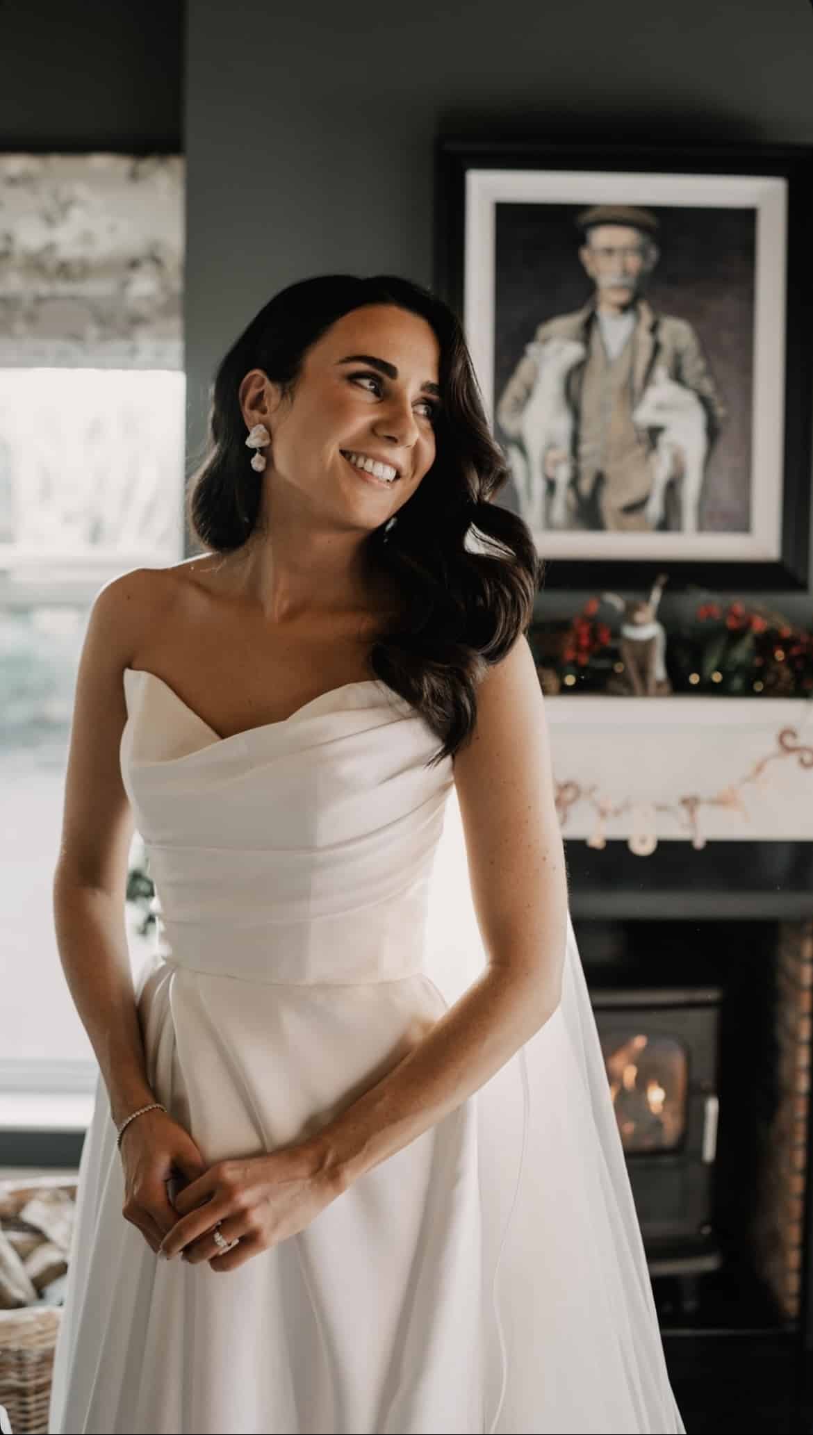 Smiling woman in a wedding dress near a fireplace with festive decor and a portrait in the background.