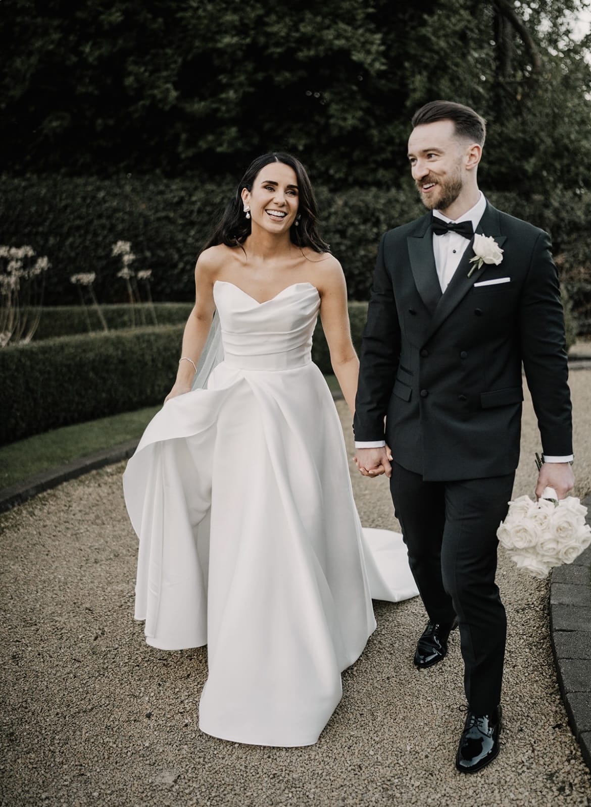 Bride and groom smiling while holding hands, walking outdoors in wedding attire.
