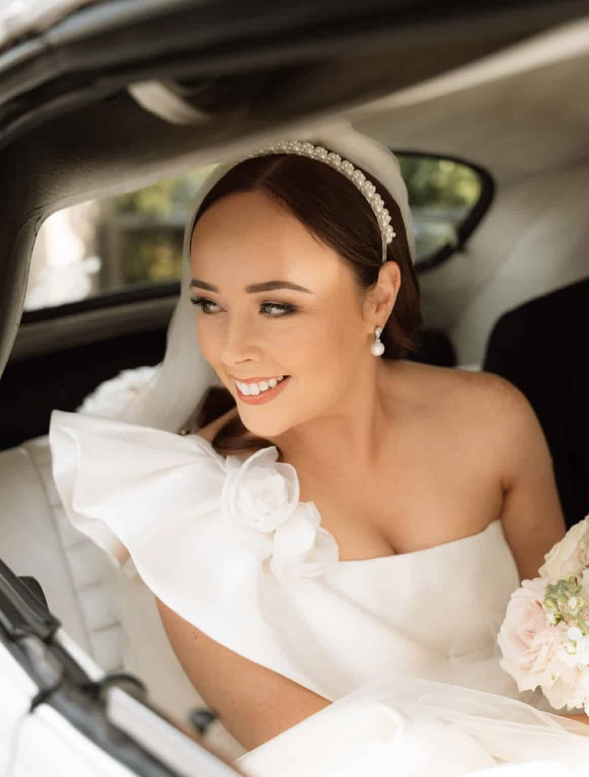 Bride smiling in car, wearing a white dress with floral detail, pearl headband, and holding a bouquet.