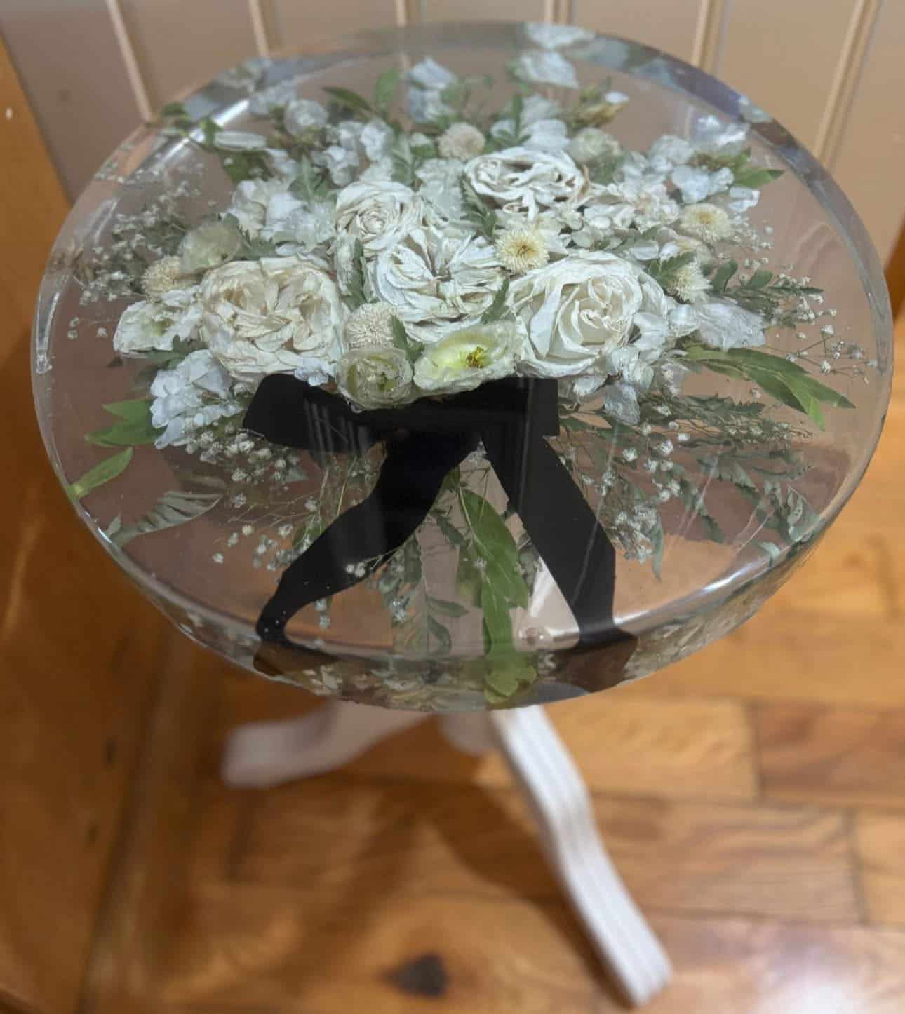 Preserved white floral bouquet with greenery under clear resin on wooden stool.