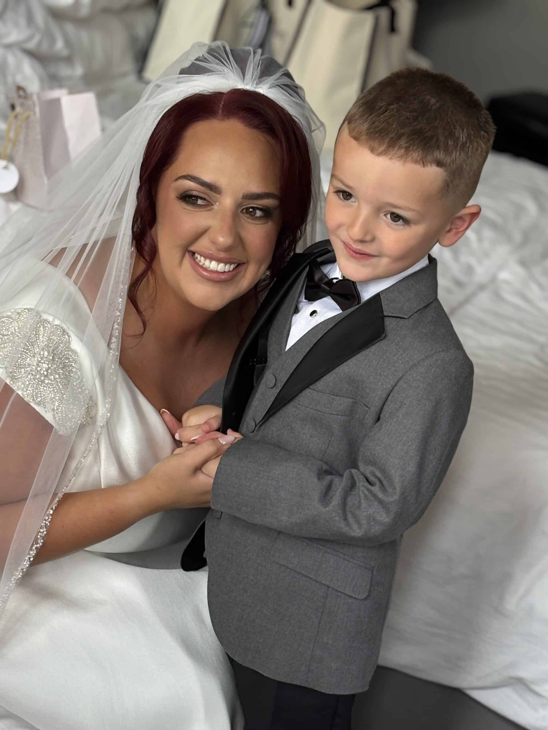 Bride smiling with a young boy in a tuxedo, holding hands and posing for a photo.