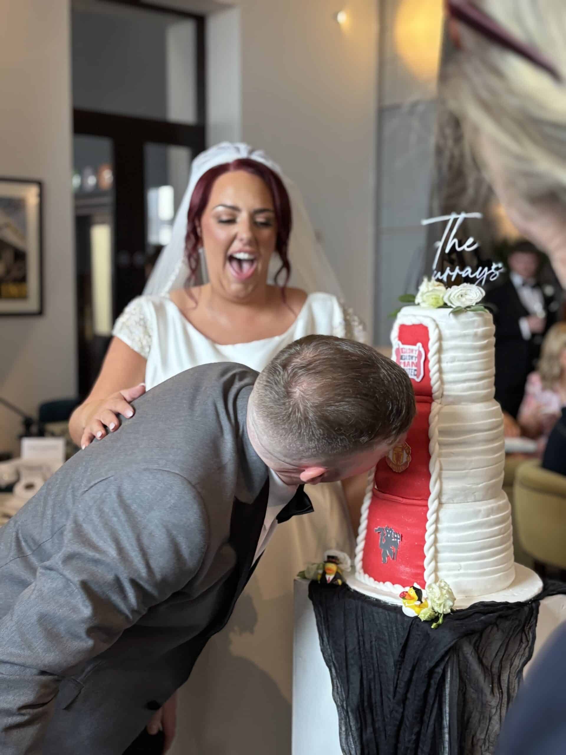 Groom playfully bites wedding cake as bride laughs, indoor setting.