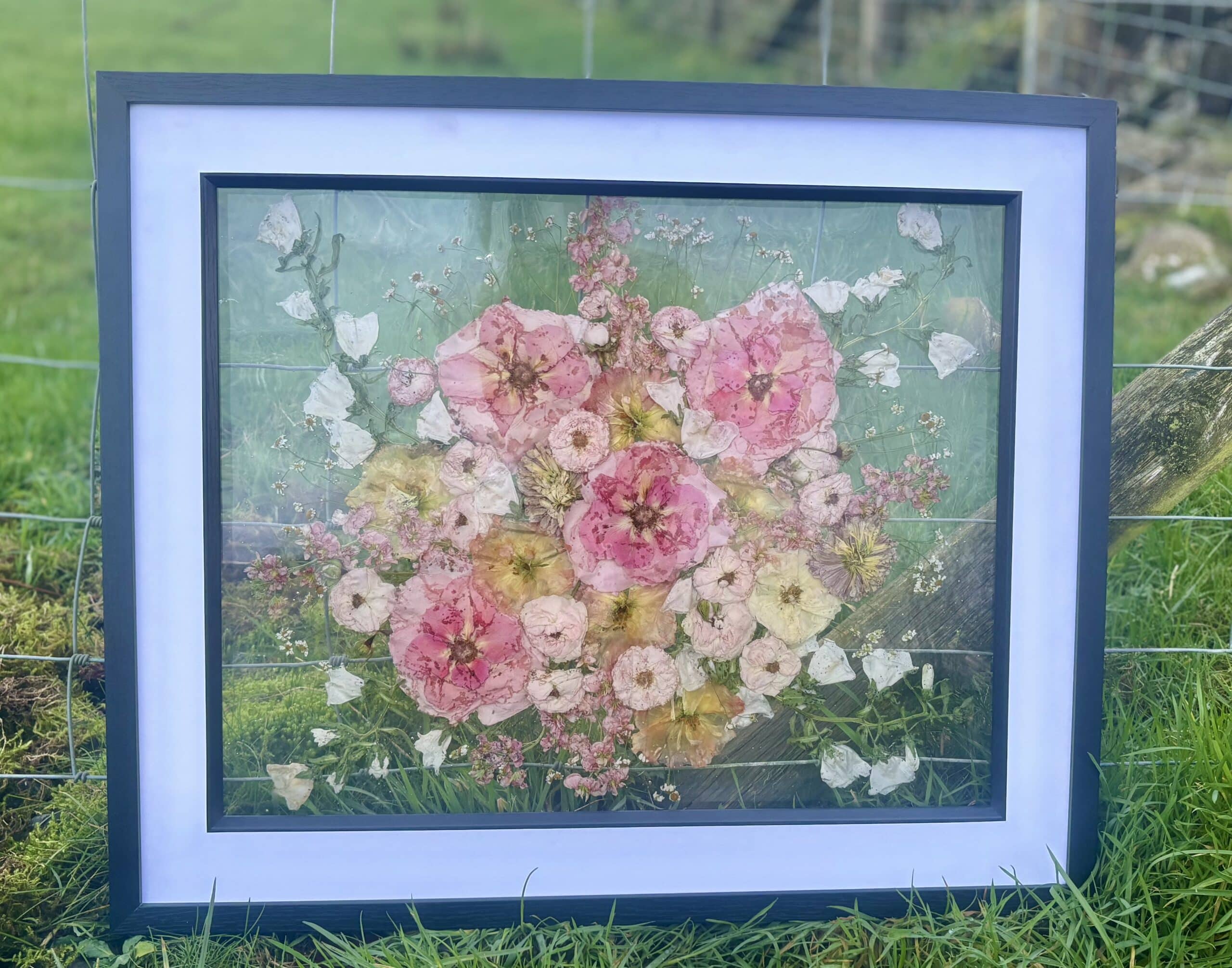 Framed floral artwork featuring pink and white flowers against a green natural background.