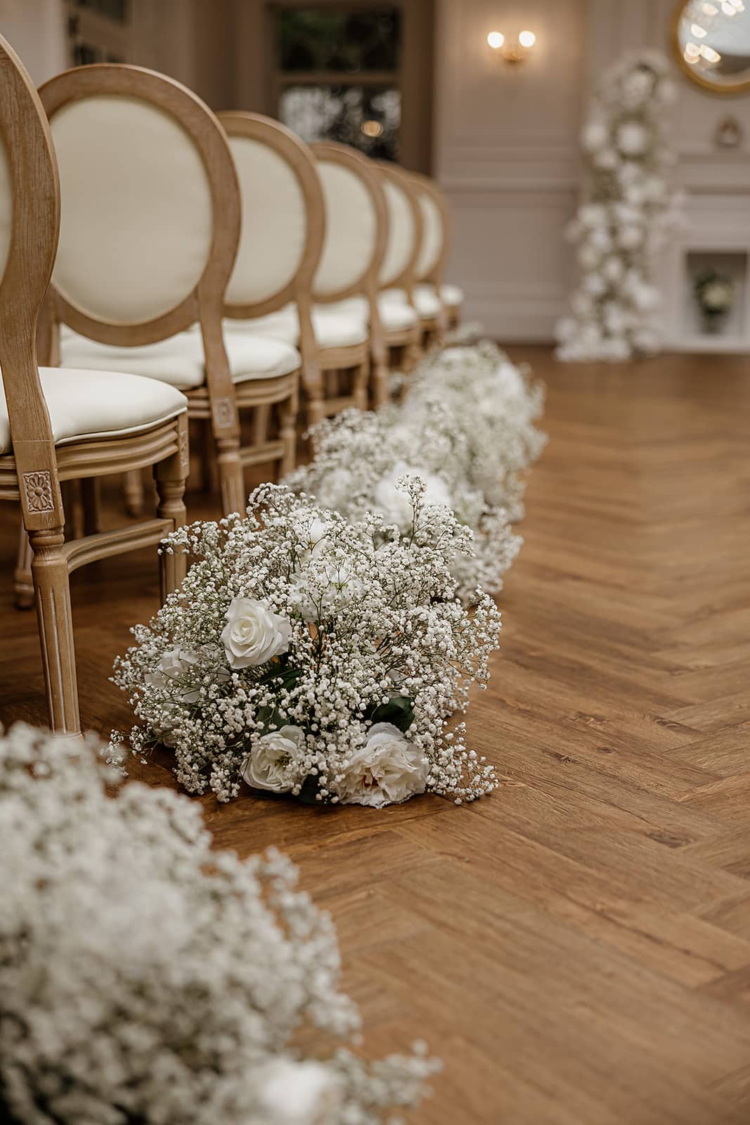 Elegant white florals lining wooden chairs on hardwood floor in a wedding ceremony setup.