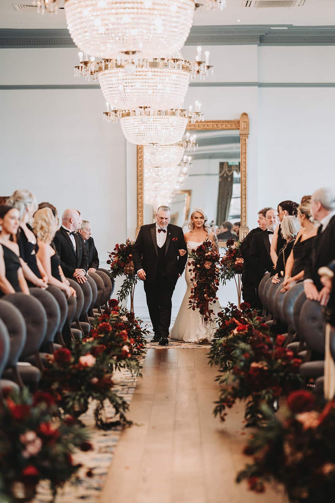 Bride walking down the aisle with escort, surrounded by guests, in an elegant venue with chandeliers and floral decorations.