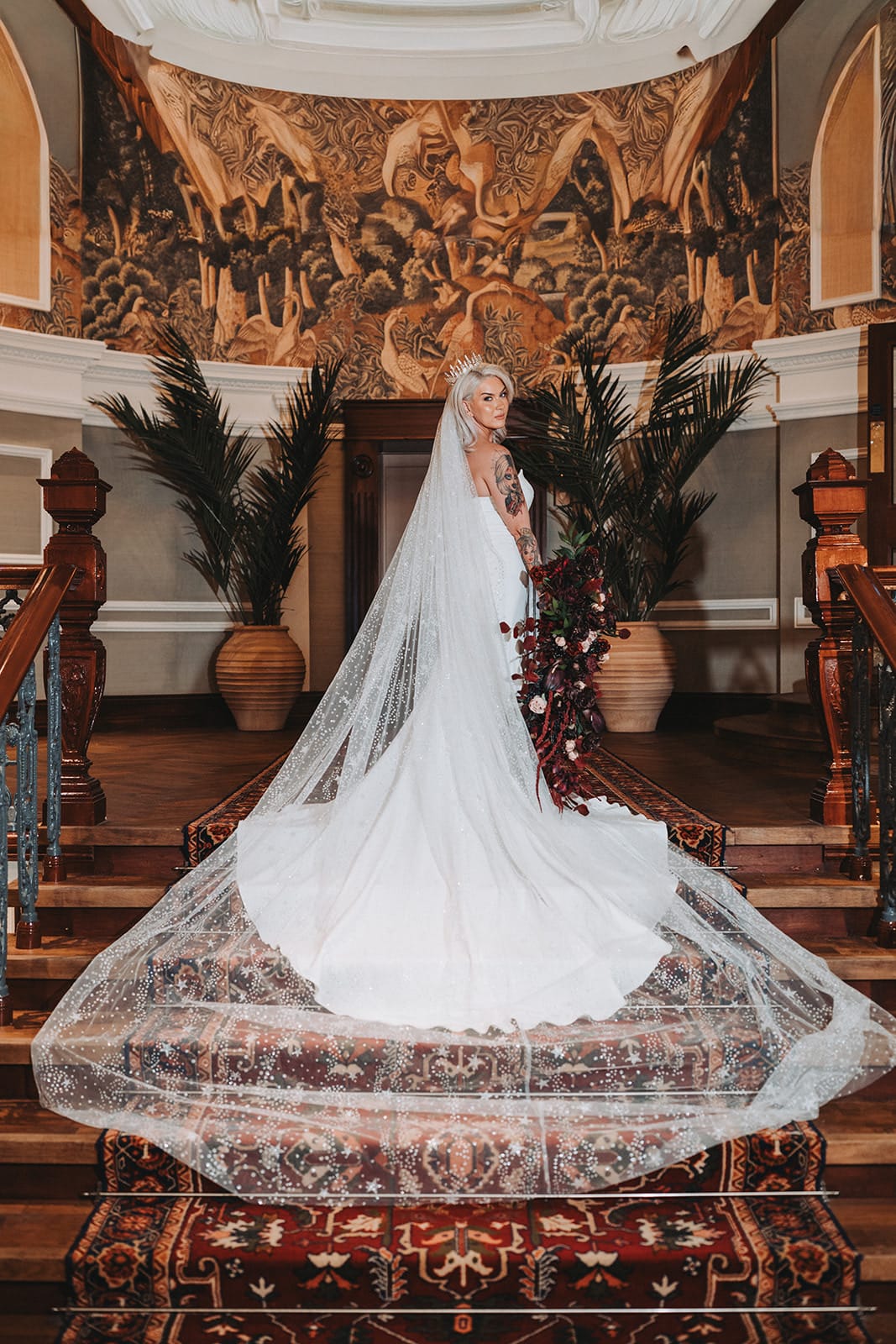 Bride in elegant gown and veil on ornate staircase with mural backdrop.
