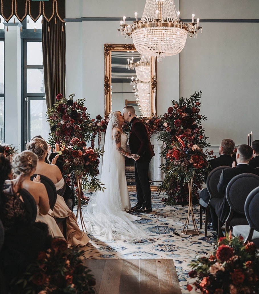 Bride and groom kissing at a wedding ceremony indoors with floral decorations and guests seated.