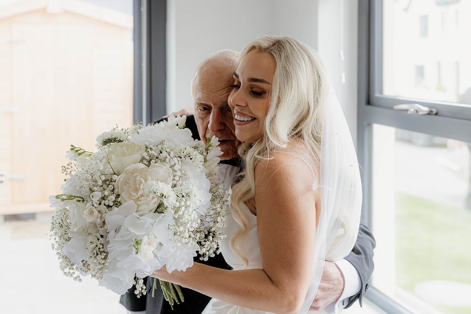 Bride hugging an elderly man, holding a bouquet of white flowers.