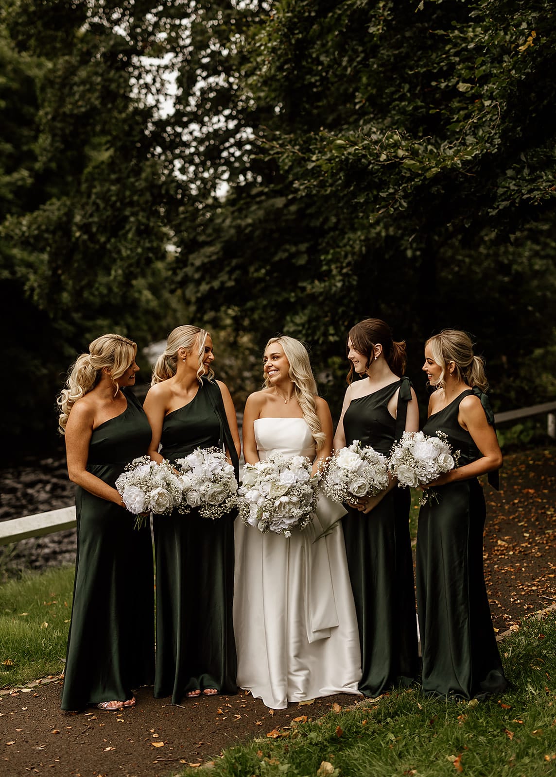 Bride and bridesmaids in dark green dresses holding white bouquets, standing outdoors on a path with green foliage.
