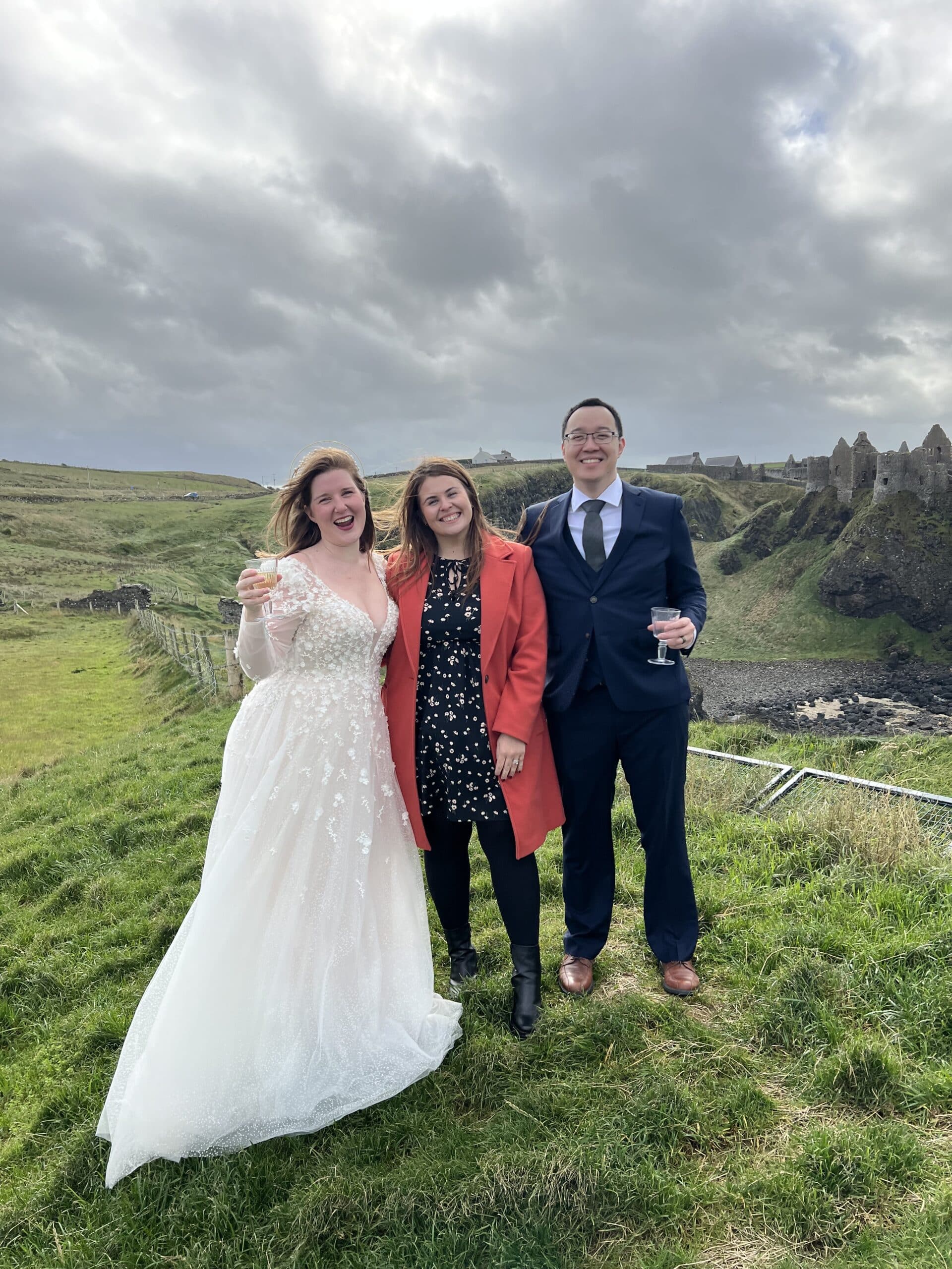 Bride with two friends on grassy hill near ruins, holding drinks, overcast sky.