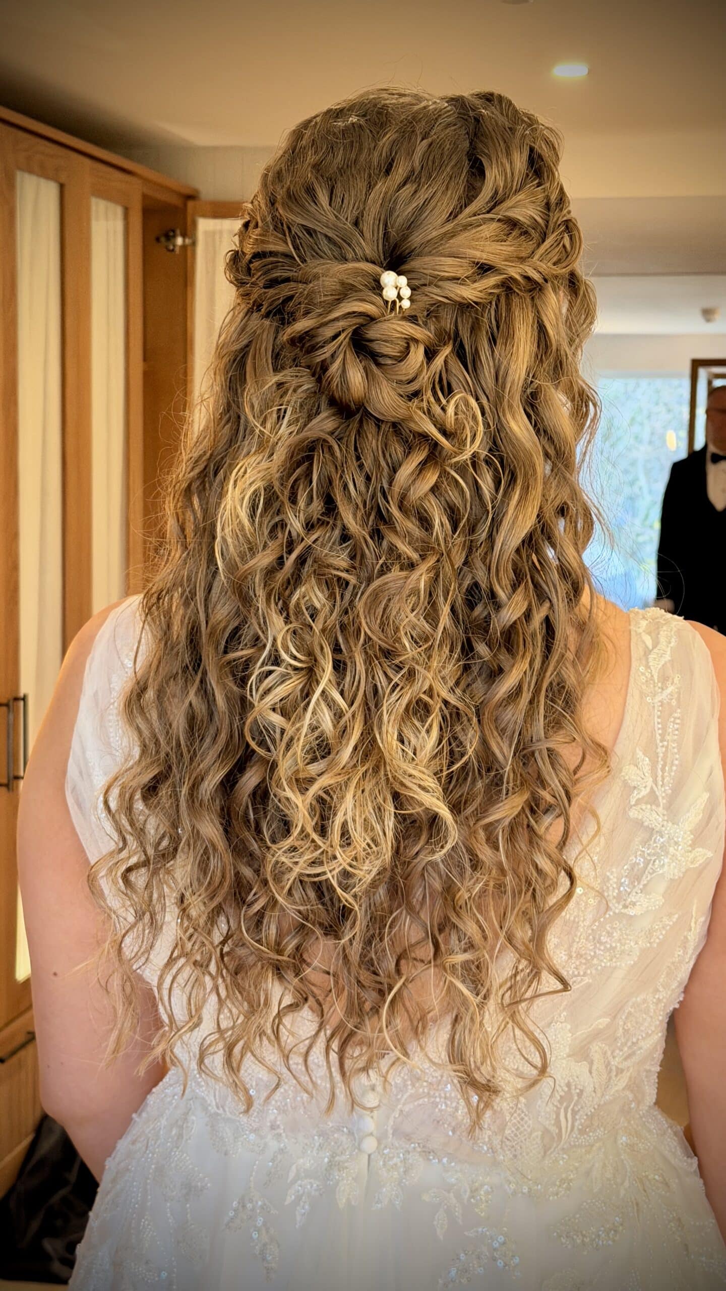 Curly bridal hairstyle with pearl hairpin, lace wedding dress, wood cabinets in background.