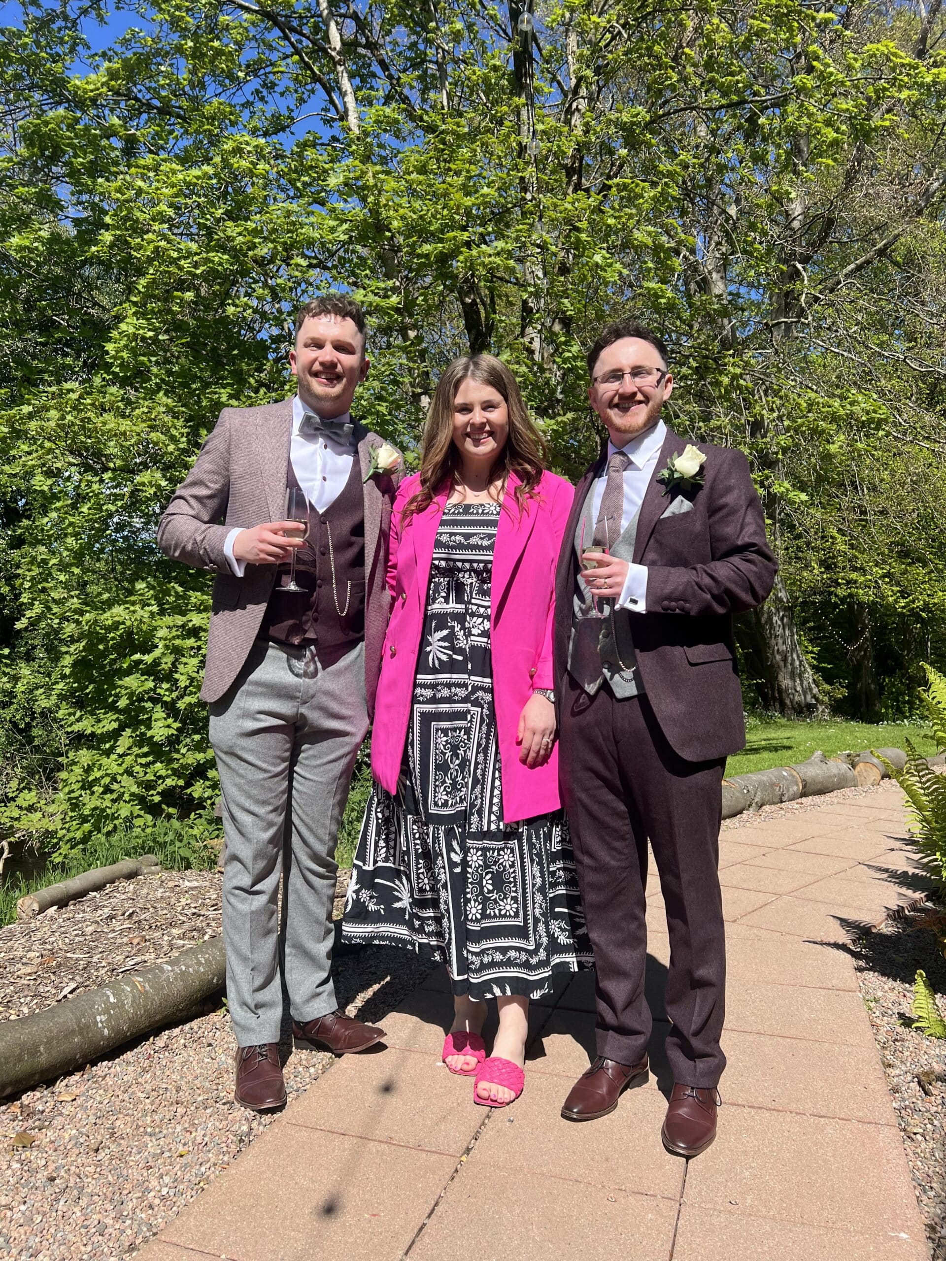 Three people dressed in formal attire standing on a garden path, with trees in the background on a sunny day.
