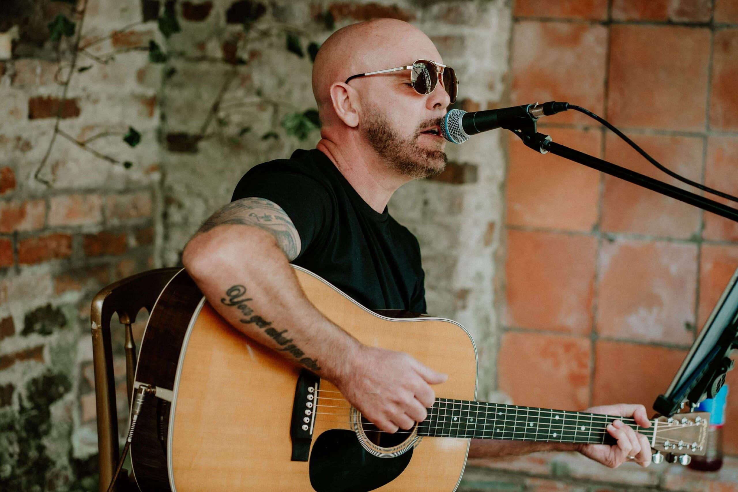 Bald man with sunglasses playing acoustic guitar and singing into a microphone.