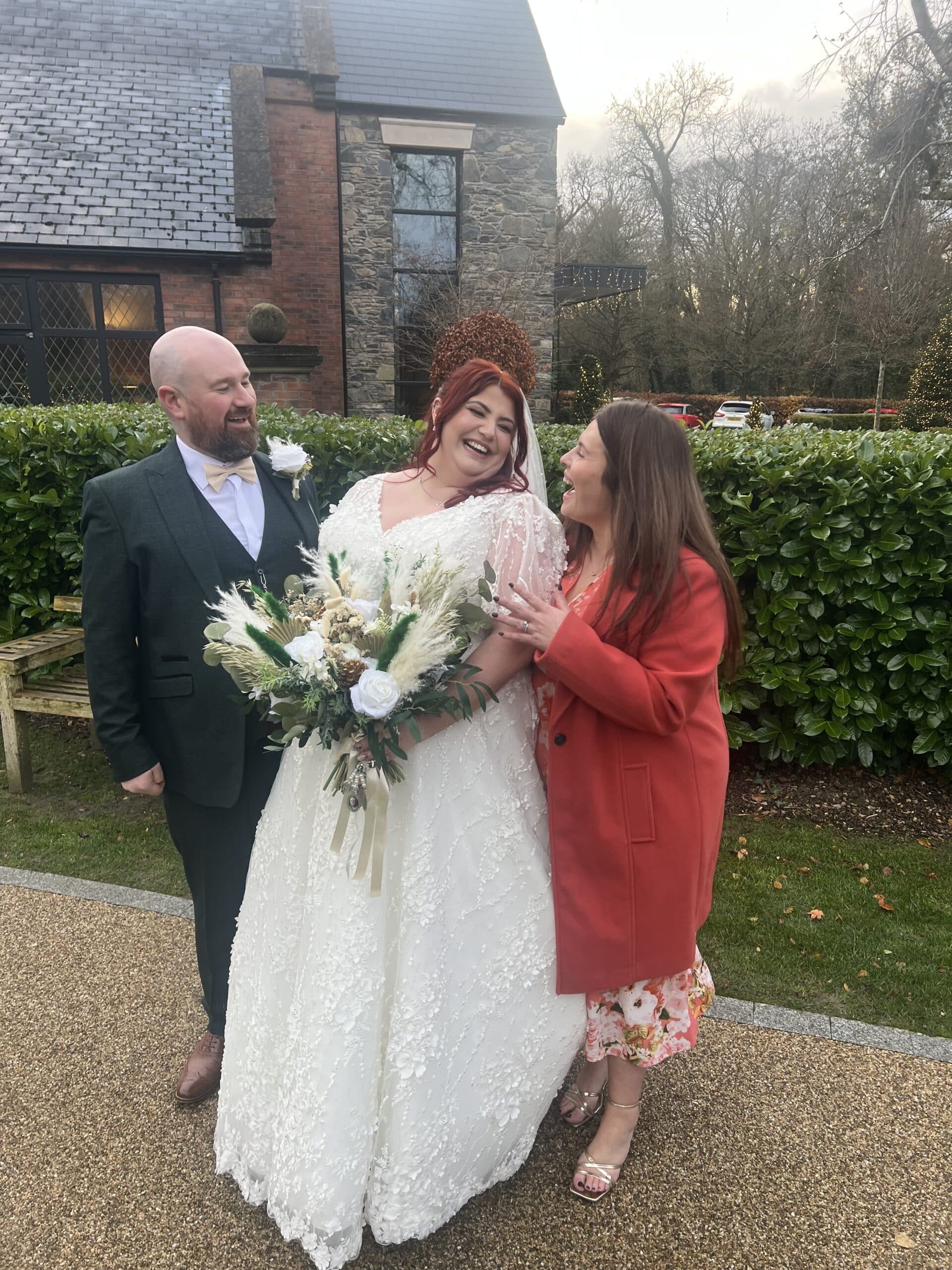 Bride in a white dress laughing with two guests outside a stone building.