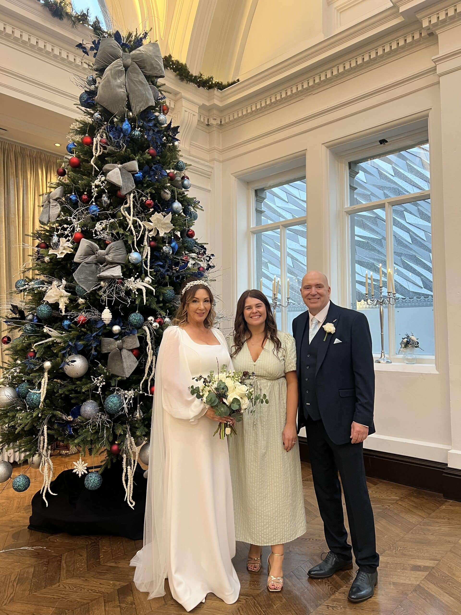 Three people pose in front of a decorated Christmas tree indoors.