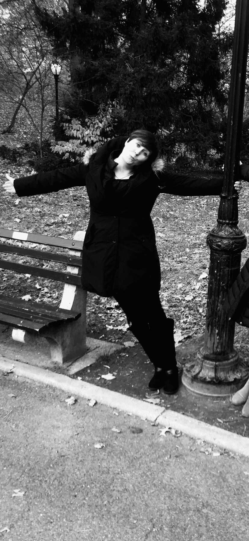 Person in a dark coat standing on a park path, arms outstretched, by a bench and lamppost, with trees in the background.