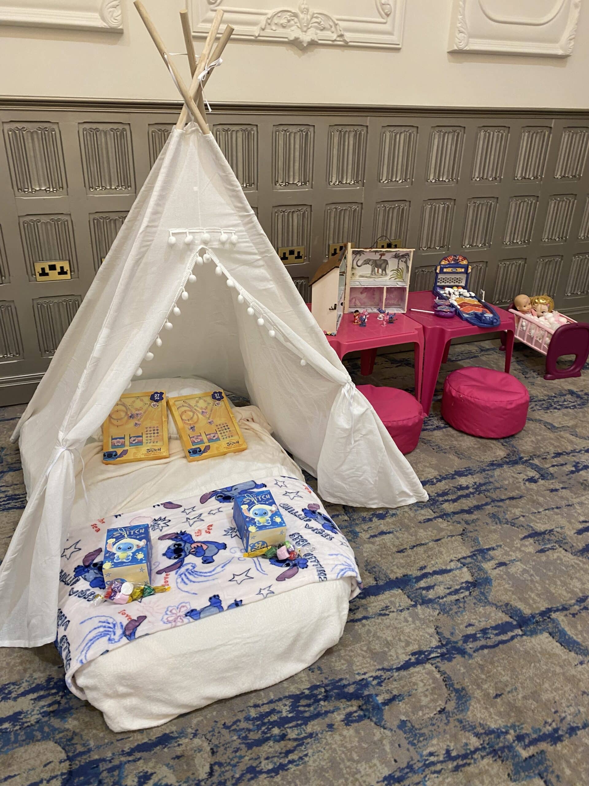 Kids' indoor play area with a white teepee tent, pink tables, cushions, toys, and a themed blanket.
