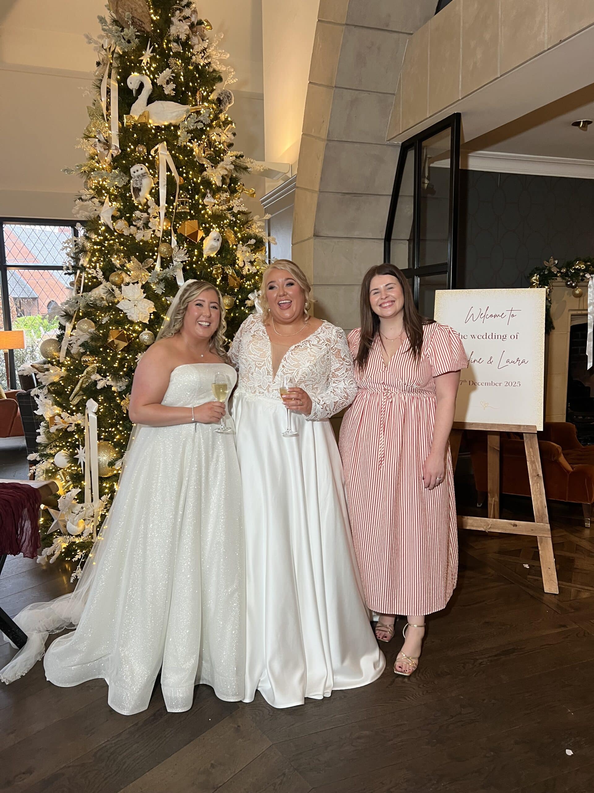 Three women in front of a decorated Christmas tree, two in wedding dresses holding champagne glasses.