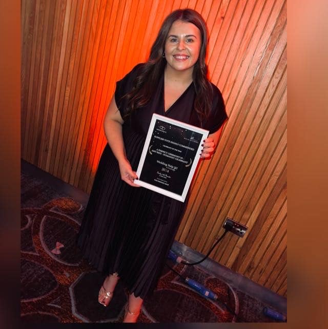 Person holding an award plaque, smiling, standing against a wooden wall.