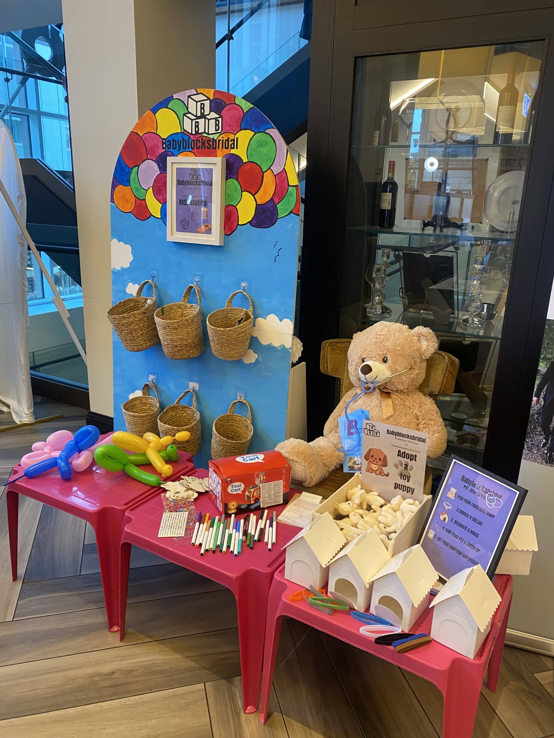 Children's craft station with colorful balloons, stuffed teddy bear, markers, toy houses, and balloons for making shapes.