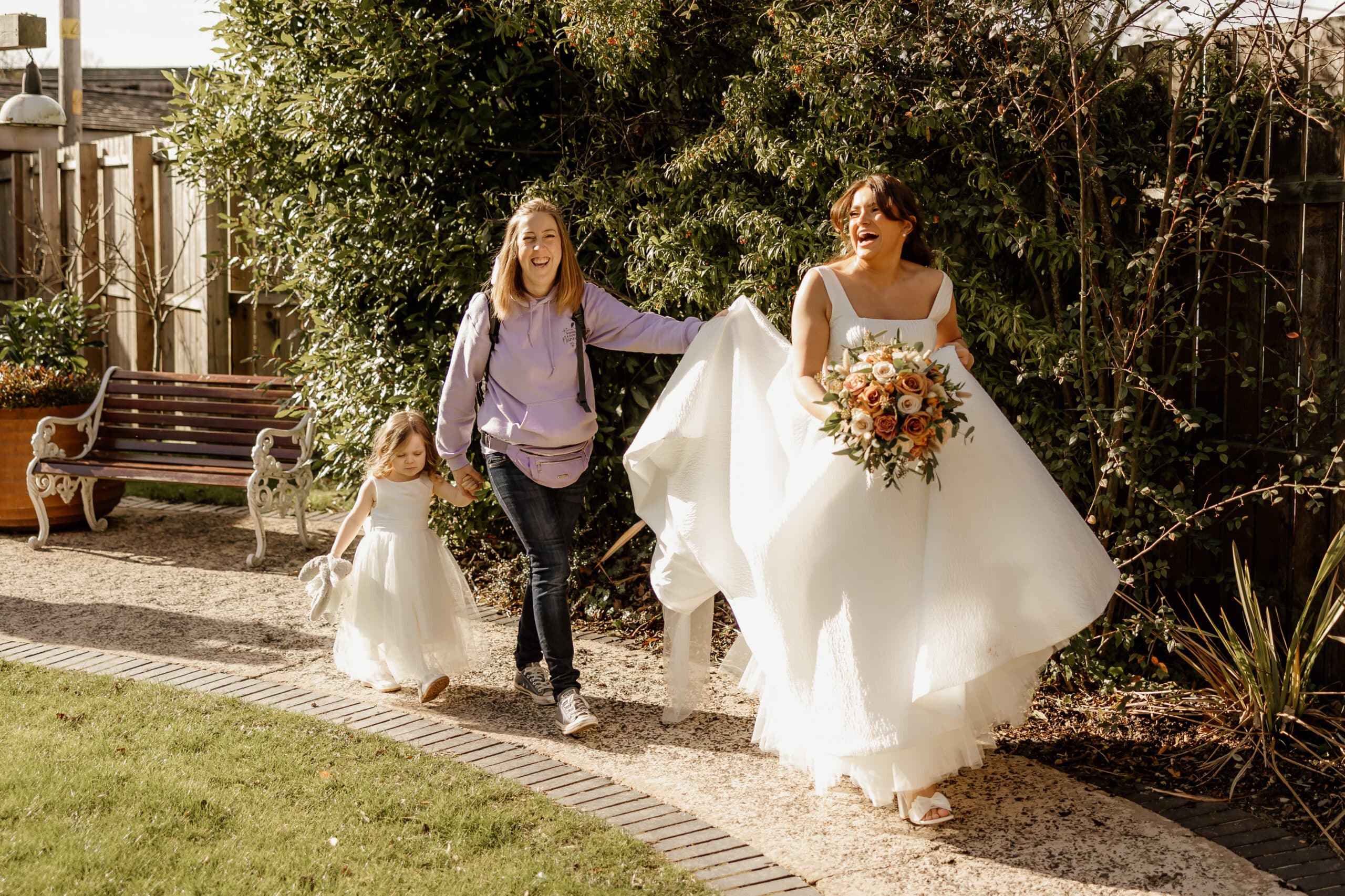 Bride with bouquet, smiling, holding dress, walks with a woman and child in white dress on garden path.