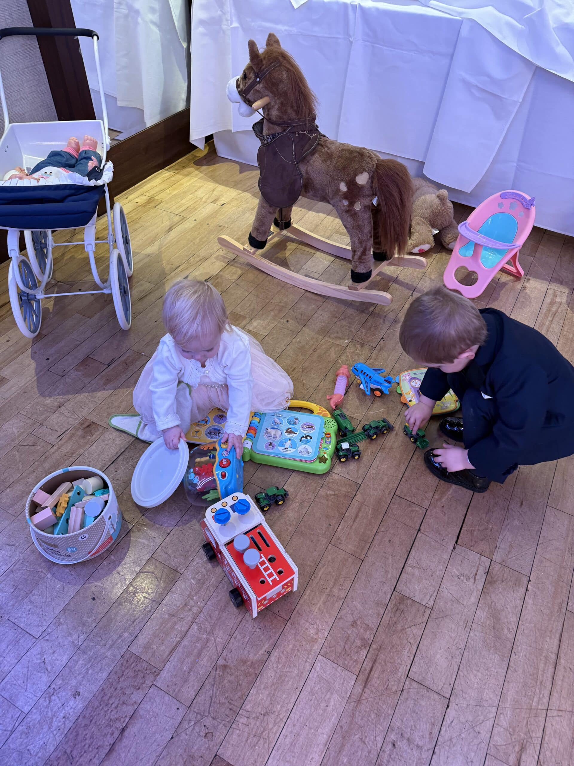 Two children playing with toys on a wooden floor, including a rocking horse, toy pram, and various toy vehicles.