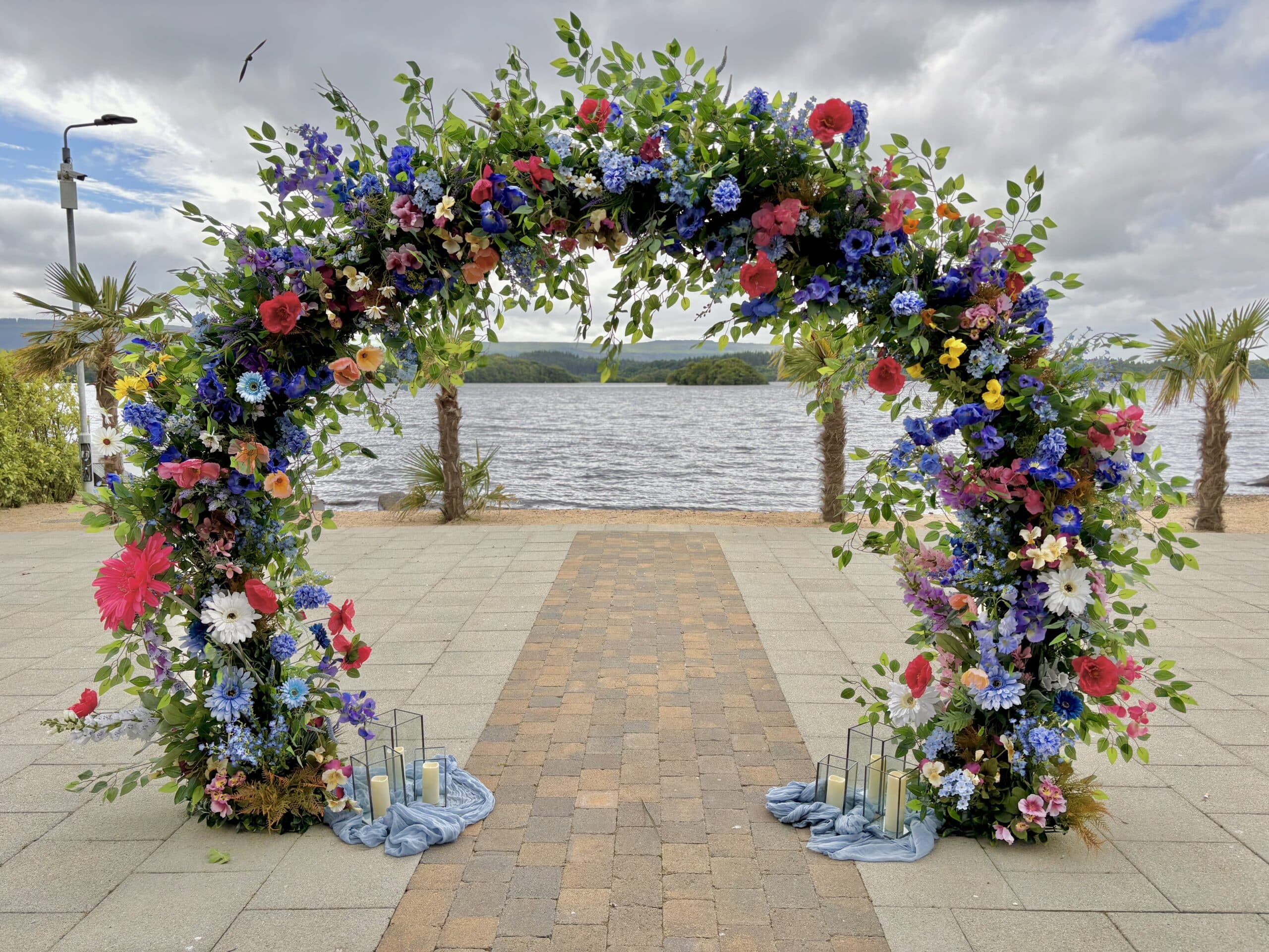 Colorful floral arch by a lakeside with overcast sky and candles at the base.