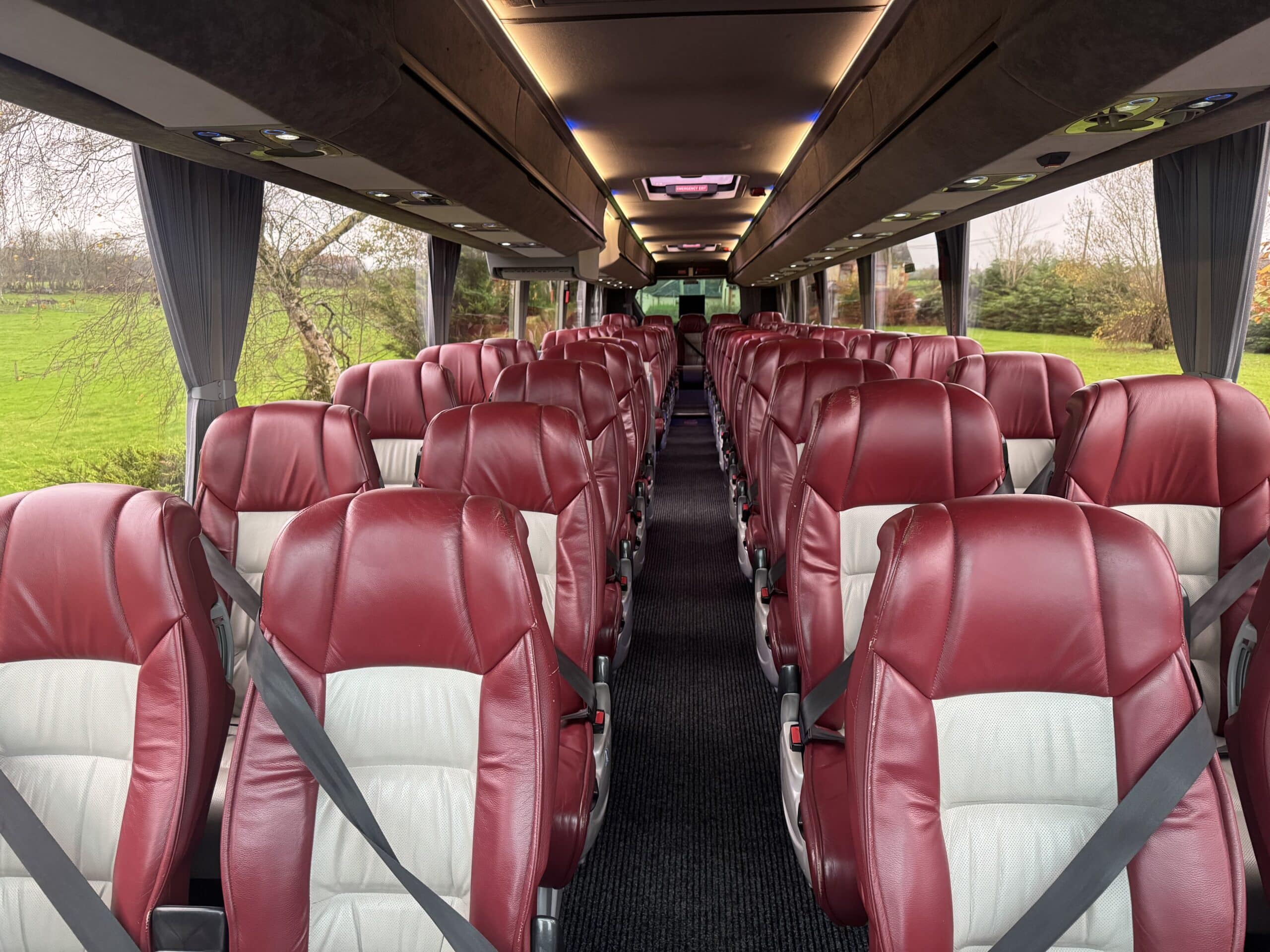 Empty coach interior with red leather seats and seatbelts, large windows showcasing greenery outside.